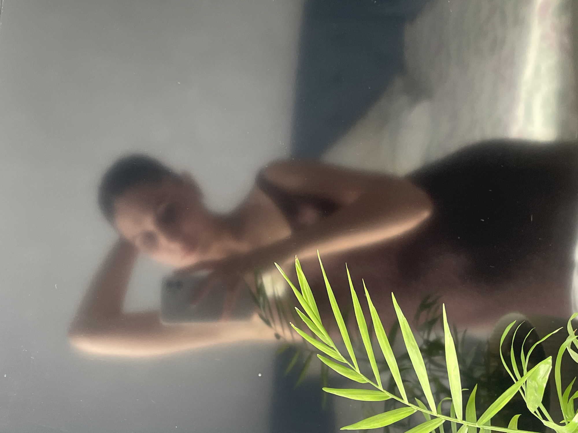 The image shows a reflection in a dark, glossy surface, possibly a television screen. A person is reflected, reclining with their head propped on their hand, holding a smartphone. A plant with green, linear leaves is situated in the foreground, partially obscuring the reflection. There is no information provided in the image to ascertain the location as Izhevsk, Russia.