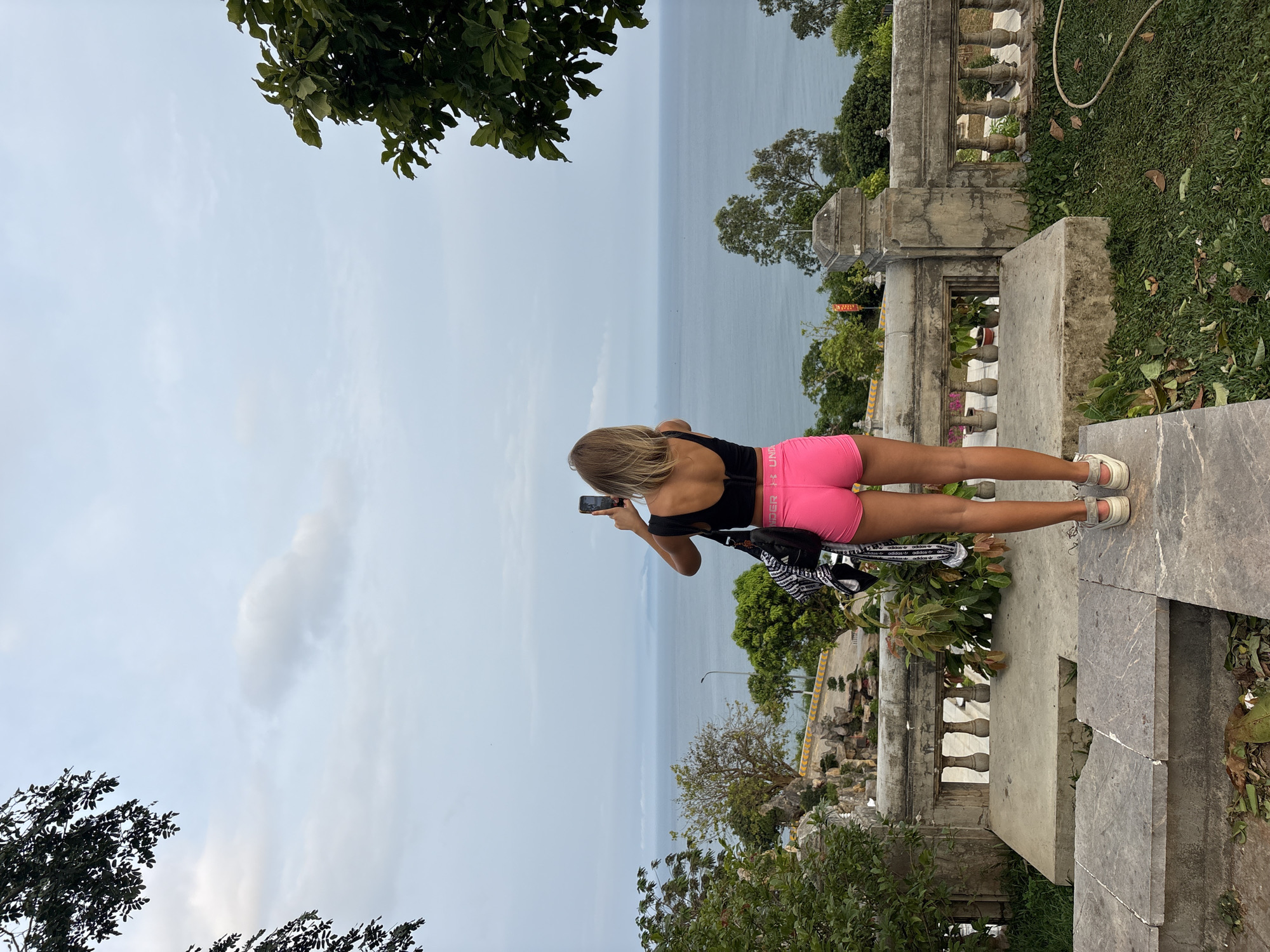 A woman stands atop a weathered stone structure, likely a historical monument or viewpoint, in Đà Nẵng, Vietnam. The location is suggested by the seaside landscape and the style of the architecture. She is positioned with her back to the viewer, facing the sea. She is clad in a black top and vibrant pink athletic shorts, and appears to be taking a photograph with her phone. The seascape stretches out before her, with trees and foliage surrounding the platform on which she stands. The sky is overcast. The platform is elevated, featuring balustrades and steps leading up to it. A white Adidas bag rests beside her. The scene conveys a sense of exploration and appreciation of the natural beauty of the Đà Nẵng coastline.