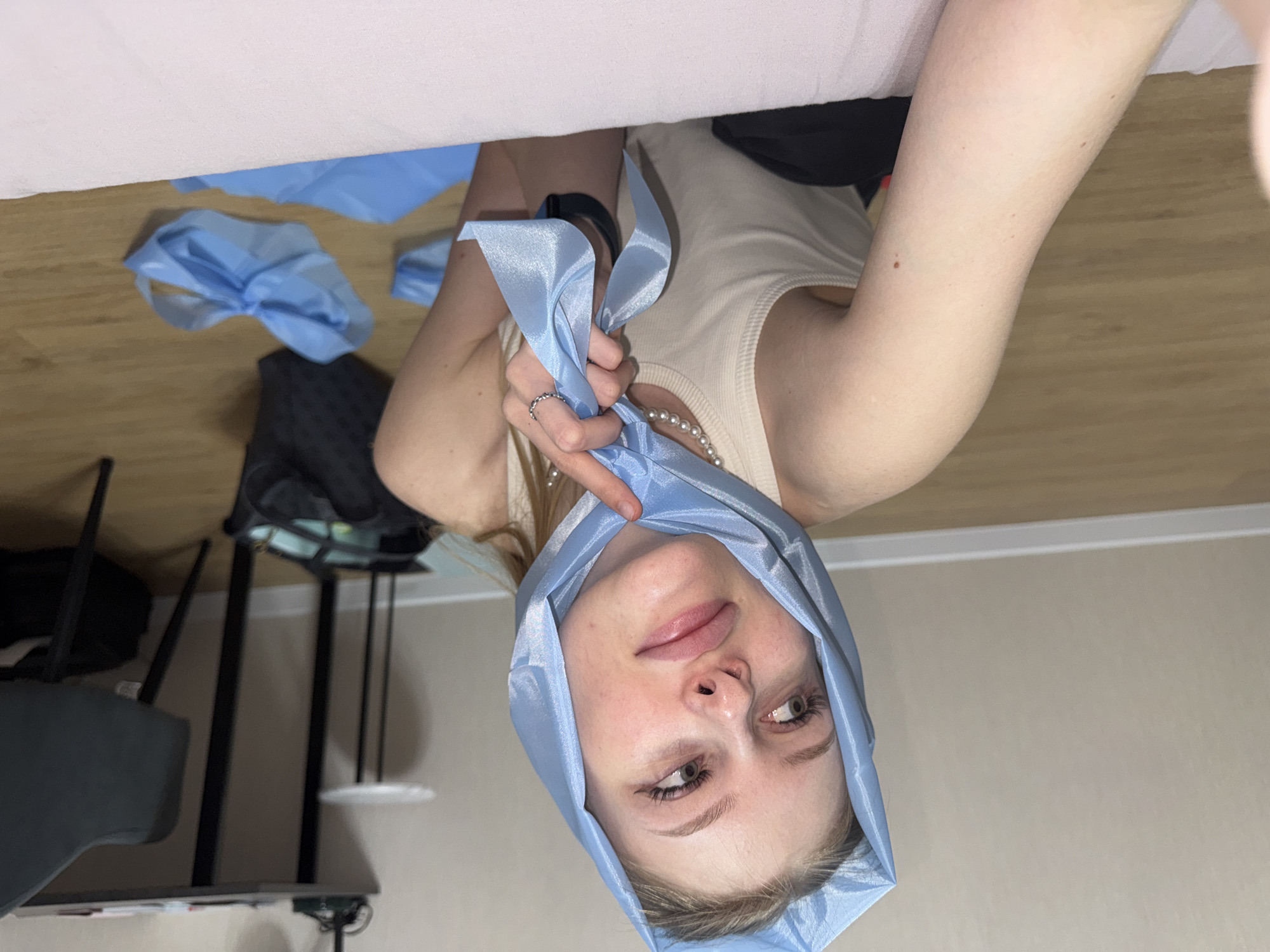 A young woman with blonde hair is looking to her left, her head covered with a light blue scarf. She is wearing a cream-colored tank top and a pearl necklace. Her left arm is bent, and her hand is holding a part of the scarf. She has on a silver ring on her left index finger. There are two blue scarves on the floor in front of her. In the background, a black desk with papers and a chair are visible, along with a small white table with a lamp on the desk. The floor is made of light brown wood planks. The location is a room in the city of Perm, Russia.
