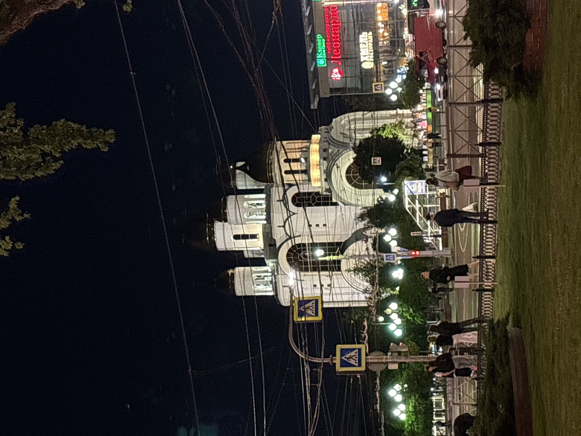 The image depicts a vibrant city scene at night in Kaliningrad, Russia, showcasing an active urban environment after dusk.
In the mid-ground, a grand, ornate white building, likely a cathedral or church, stands prominently with multiple domes and arched windows, brightly illuminated against the dark sky. A complex network of power lines stretches across the upper left portion of the frame.
To the left of the cathedral, a street is visible with vehicles, including a red truck, and several commercial buildings. These buildings feature illuminated signs with Russian text. Visible signs include "Клевер" (Klever), identified as a cash & carry store; "Леонардо" (Leonardo), a "Хобби-гипермаркет" (Hobby-hypermarket); and "ХМЕЛЬ РЕСТОРАНЪ" (Khmel Restaurant).
In the foreground, a well-maintained grassy area occupies the lower right, separated from a pedestrian path by a decorative metal fence. Several people are seen walking along this path, some casually strolling. Streetlights cast bright beams, illuminating parts of the path, people, and surrounding trees. Yellow pedestrian crossing signs are also visible, indicating designated safe zones. The sky is dark, confirming the late evening or night, under clear weather conditions.