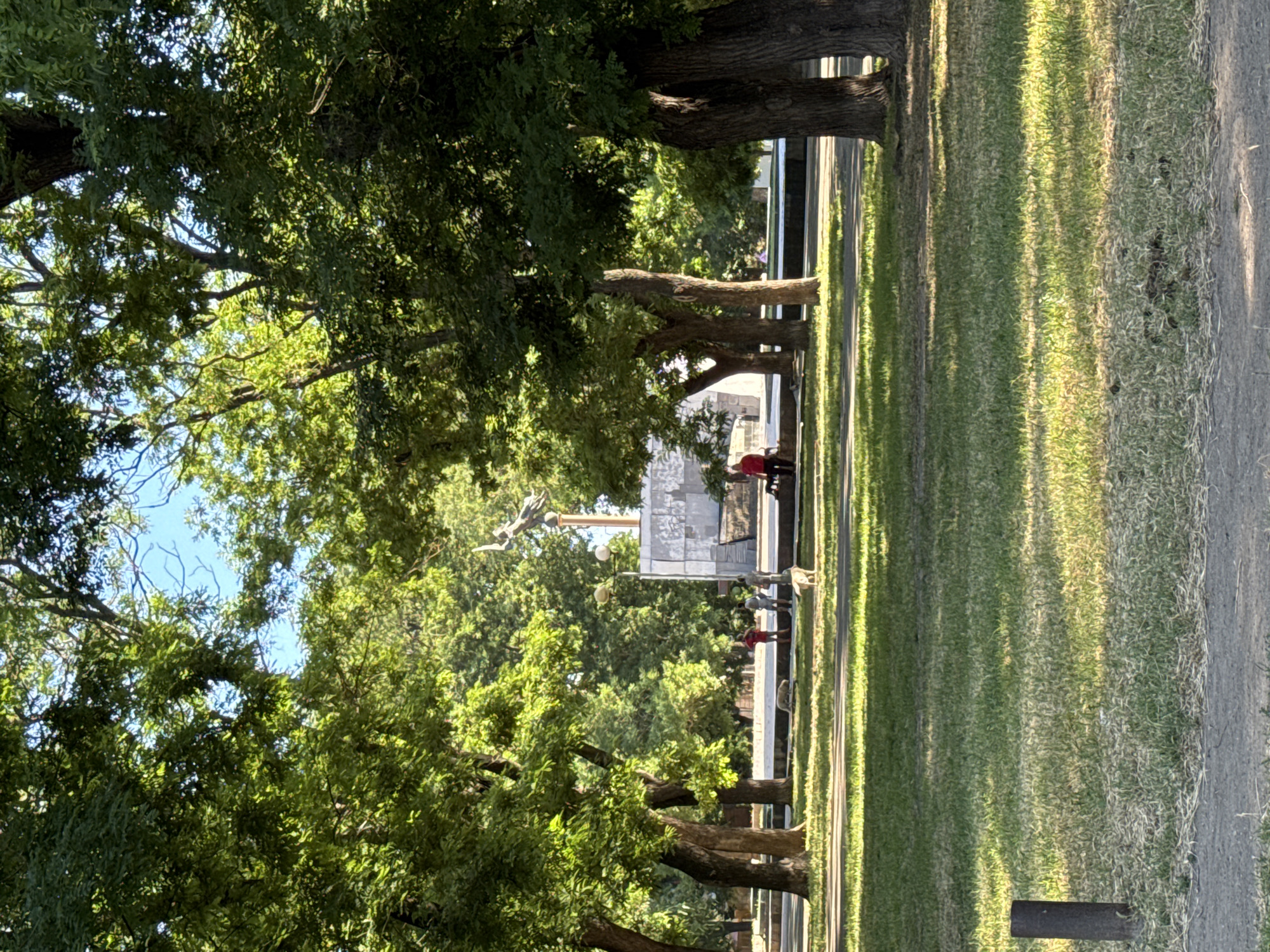 The image depicts a public park in Odesa, Ukraine, characterized by mature trees with dense green foliage and a well-maintained lawn. In the background, a monument featuring a sculpted figure atop a pedestal is visible. Several individuals are present in the park, some walking or standing near the monument. Two dogs are also present on the lawn, one appearing to be a light-colored breed. The scene is bathed in sunlight, casting dappled shadows across the grass. A low, dark cylindrical object, possibly a bollard or post, is situated on the lower left side of the frame, adjacent to a paved walkway. The overall atmosphere suggests a tranquil, everyday scene in a city park.