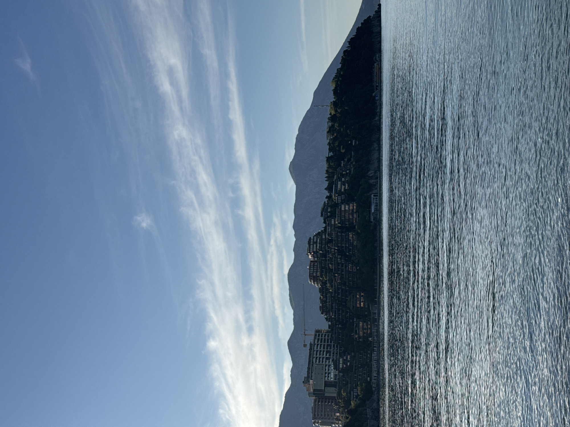 The image shows a view of the Adriatic Sea in Budva, Montenegro, with mountains in the background. The sky is blue with wispy clouds. The water is rippled. A mountainside populated with buildings, including high rises, is on the coast, and a crane is visible in the construction area.