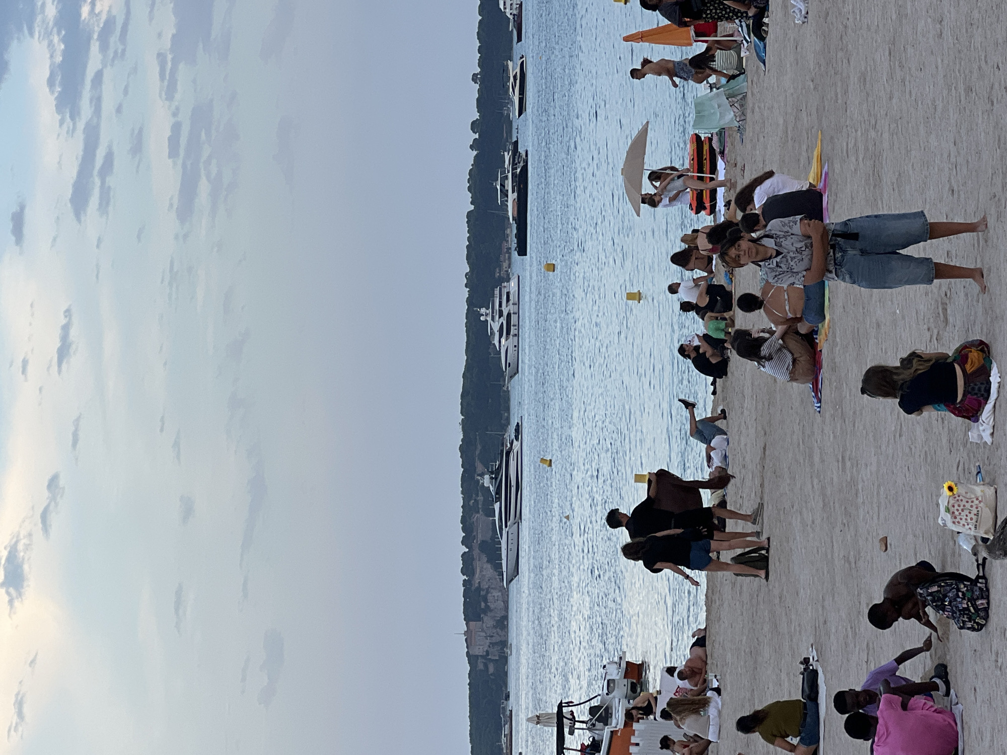 The image depicts a busy beach scene in Cannes, France, with numerous people enjoying the water and sand. In the foreground, several individuals are seated on towels and blankets, some interacting with each other. A woman with short brown hair and glasses stands in the center, arms crossed, wearing a patterned short-sleeved shirt and denim shorts. Further back, more beachgoers are visible, including a couple standing near the water's edge and a group gathered under a beach umbrella. The background features a body of water with several yachts and boats sailing, and a coastline with buildings and lush vegetation. The sky is partly cloudy, with soft, diffused light suggesting it might be late afternoon or early evening. The overall atmosphere is relaxed and summery, characteristic of a popular tourist destination like Cannes.