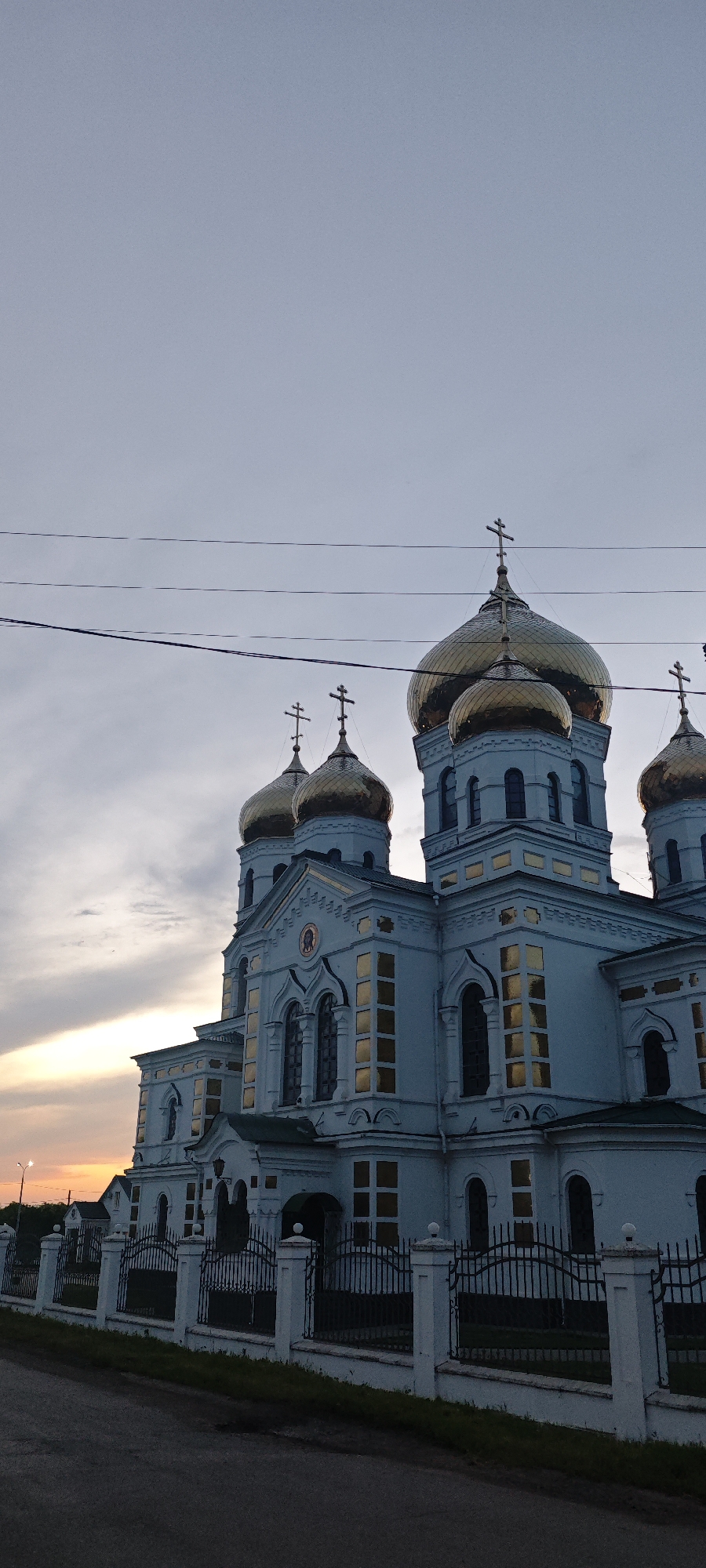 The image shows a large, ornate church with golden domes, likely an Orthodox cathedral, in Novodonetskaya, Russia. The building is light blue with gold accents, visible in the windows and trim. It has multiple tiers and several prominent golden domes with crosses on top. The sky is overcast, with a faint sunset visible on the left horizon. A fence runs along the bottom of the frame, separating the church from the road. Power lines are visible in the sky above the church. The overall scene is of a twilight setting with the church as the dominant subject.