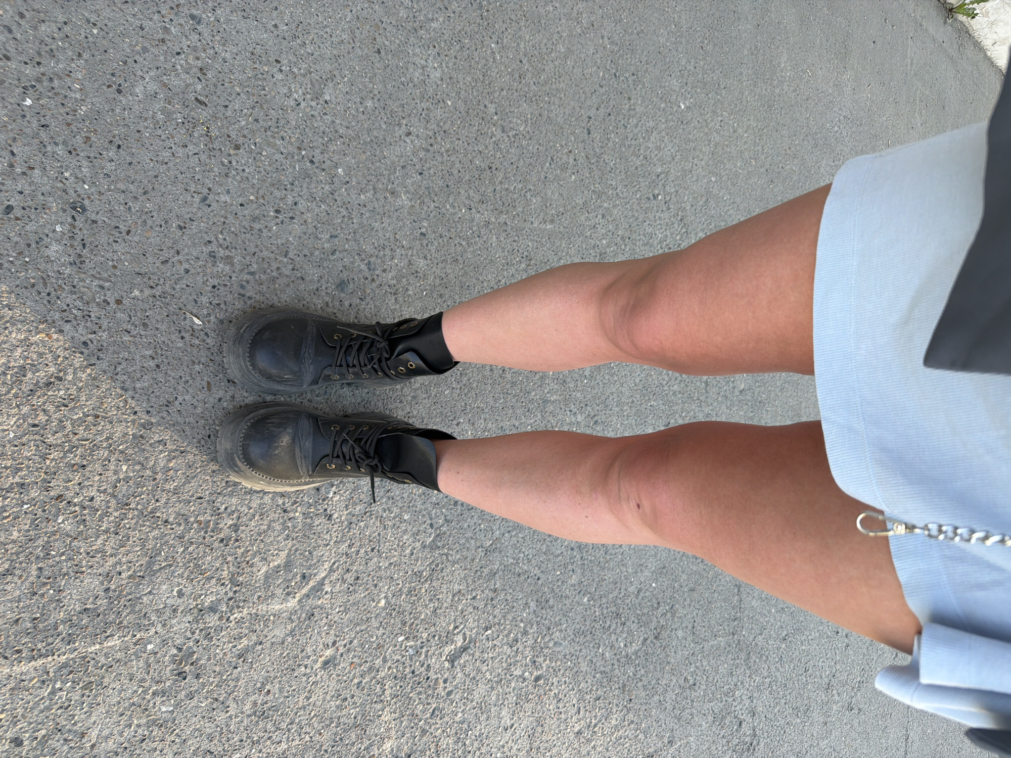A top-down shot depicts a person standing on a gray concrete surface. The person is wearing black lace-up boots. Their legs are bare from the boots to the bottom edge of a light blue garment, likely a skirt or shorts. A silver chain with a clasp is attached to the garment. The location appears to be outdoors, possibly a sidewalk or paved area. The scene is sunlit. It is not possible to ascertain that the image was taken in the city of Chaikovsky, Russia.