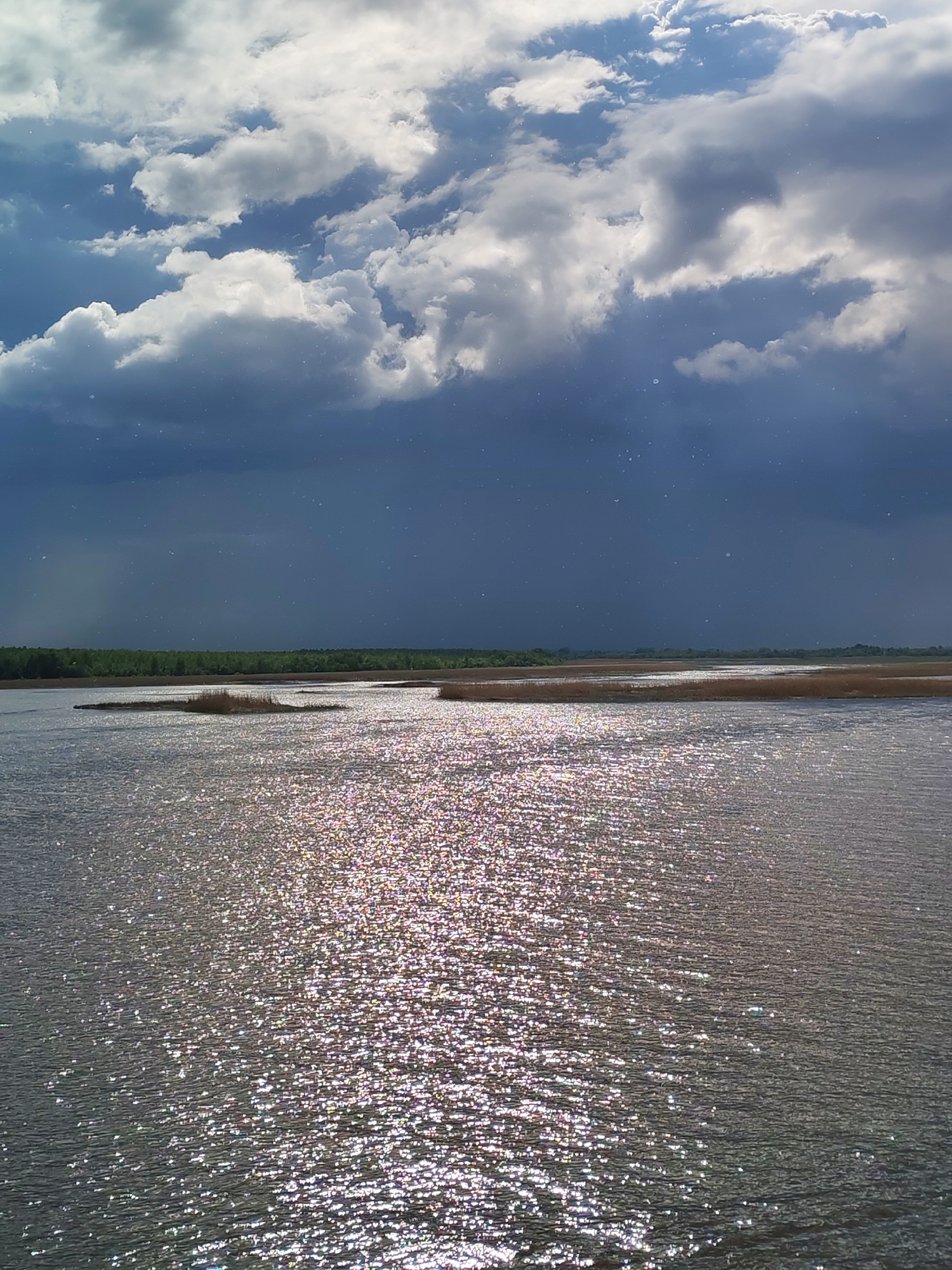 The image captures a body of water under a dramatic sky. A mix of white and gray clouds dominate the upper portion of the image, with darker, rain-laden clouds suggesting a possible storm. Light reflects off the water's surface, creating a shimmering effect. In the distance, there's a line of trees and vegetation, possibly indicating the shoreline of the water body. Given the context of Krokhino, Russia, this could be a view of the Sheksna River or a nearby lake or reservoir.