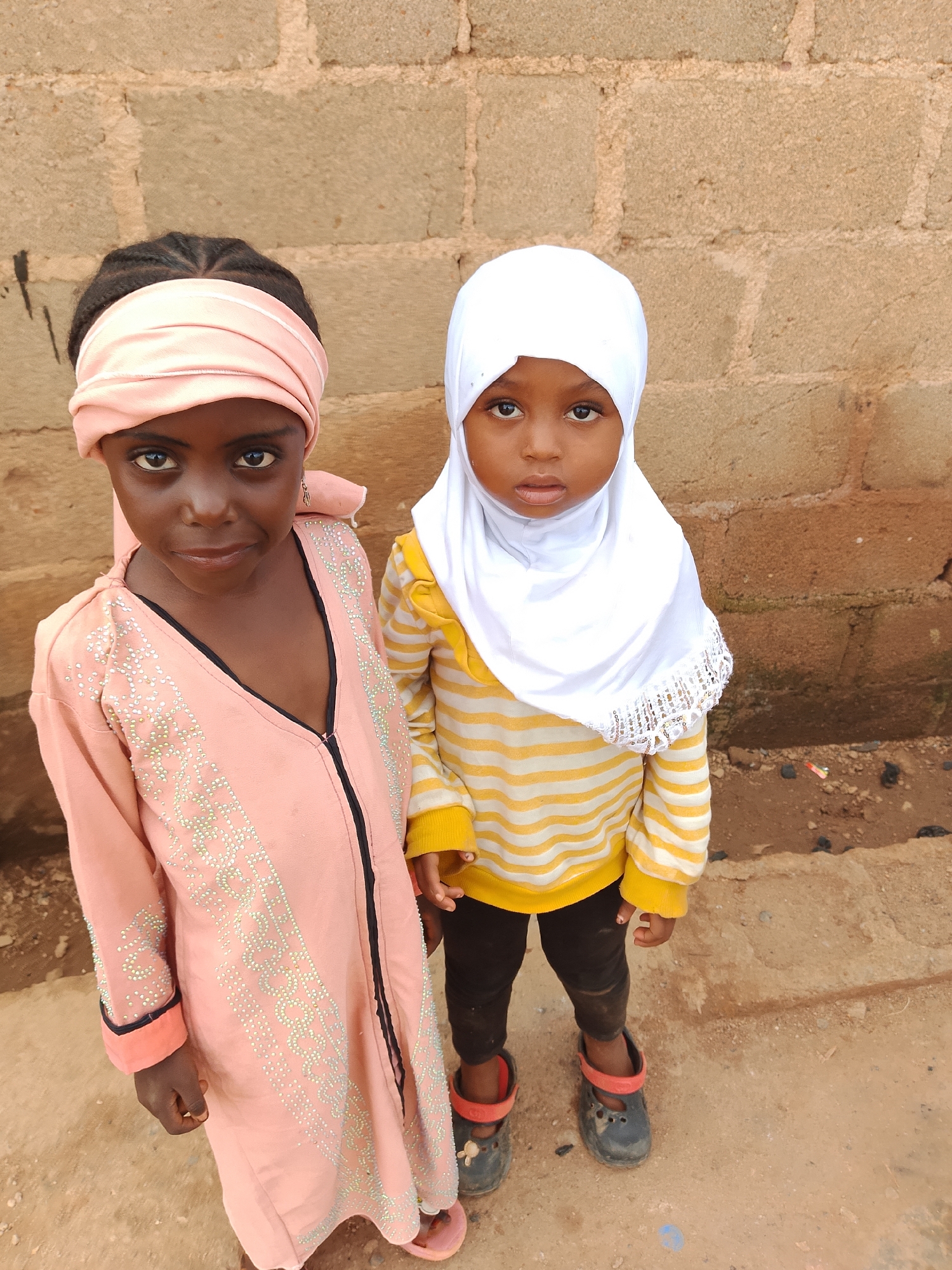 Two young Black girls stand next to each other in front of a brick wall. The girl on the left wears a pink headscarf and a peach-colored abaya adorned with rhinestones. The girl on the right wears a white hijab and a yellow and white striped long-sleeved shirt with black pants and dark clogs with red straps. The ground beneath them is dirt.