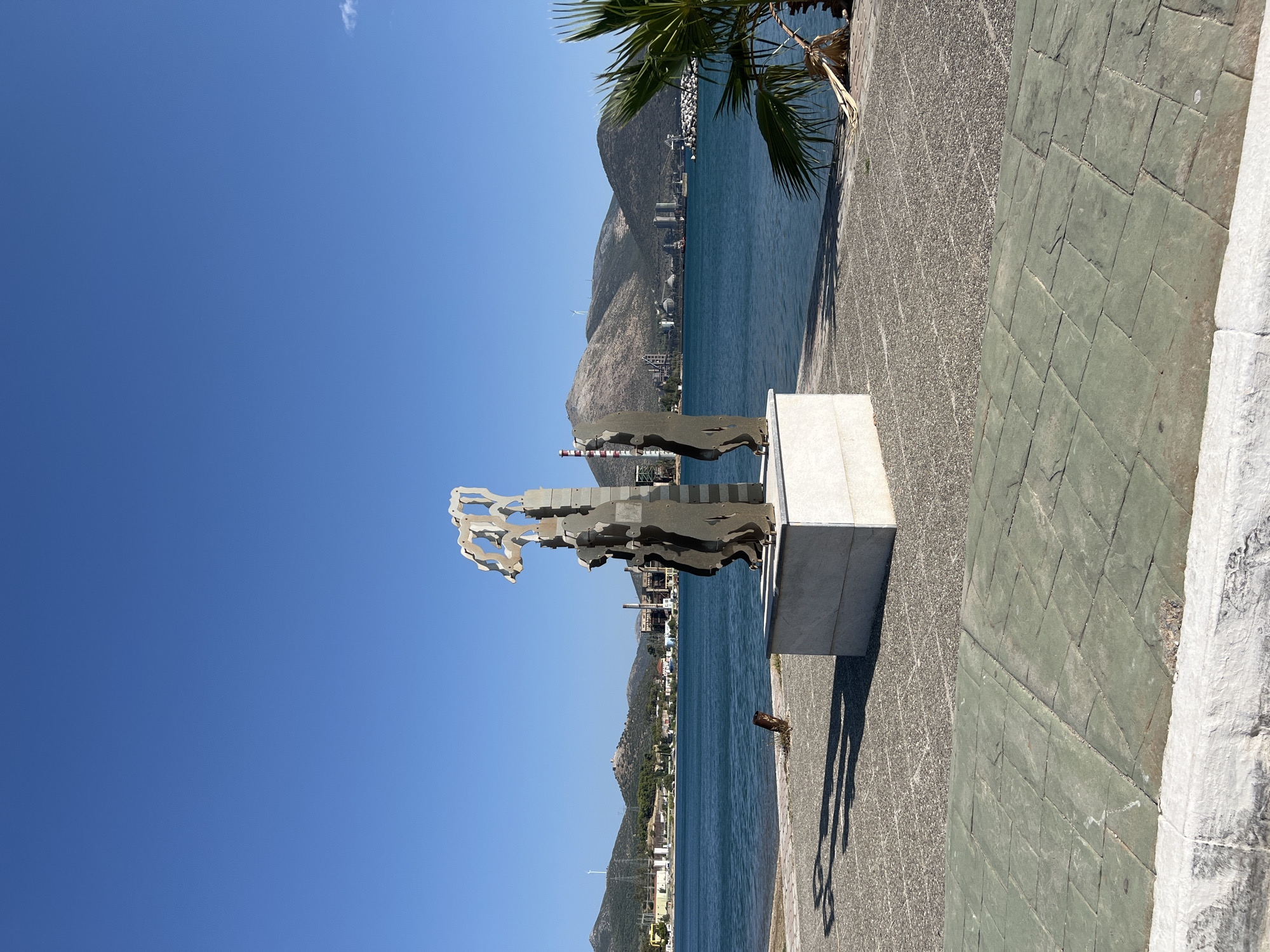 An outdoor, eye-level, medium shot shows a modern art sculpture standing on a pedestal in the foreground, against a backdrop of the sea and a distant industrial coastline under a clear blue sky. The sculpture, made of dark, metallic material, appears to be abstract figures standing together. It is situated on a paved area with small grey stones, adjacent to a walkway with larger, green-tiled paving. A white, textured curb borders the foreground. To the right, a palm frond is visible, casting a shadow. In the mid-ground, the teal-colored sea stretches out, meeting a hilly coastline dotted with industrial buildings, smokestacks, and wind turbines. The scene is located in Karavos, Greece.