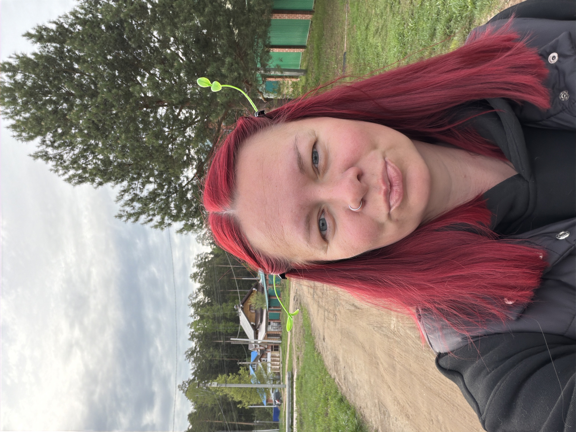 The image is a selfie of a woman with red hair, wearing a black jacket. She has a nose ring and green accessories in her hair. Behind her, a dirt road stretches into the distance. There are trees on both sides of the road, with a building visible in the background, typical of a rural area. Based on the location of Chaikovsky, Russia, it seems to be a rural location on the outskirts of the city.