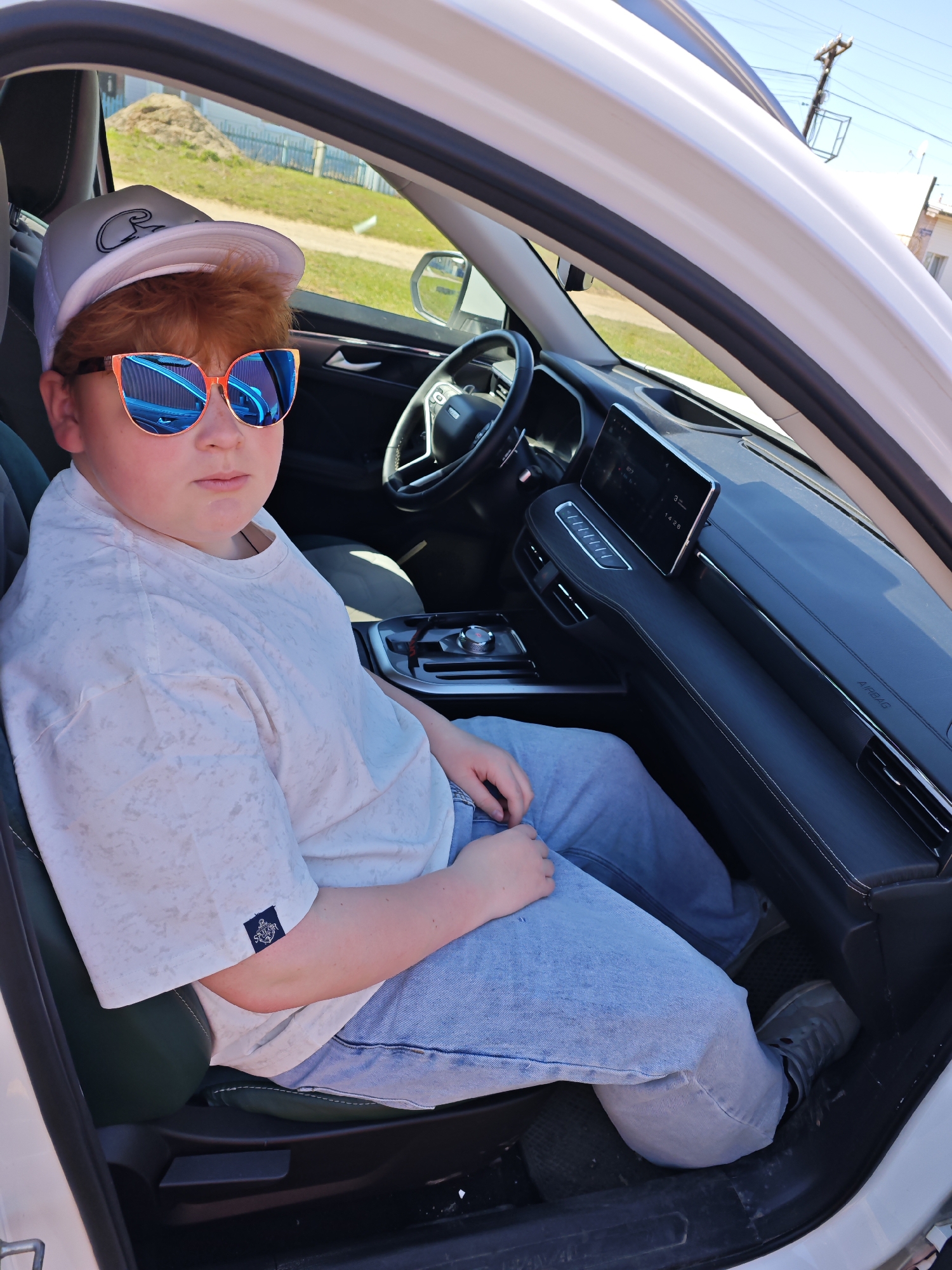 A young person with red hair and sunglasses sits in the driver's seat of a white vehicle. The individual is wearing a light-colored t-shirt, blue jeans, and a white baseball cap. The interior of the car features a black dashboard with a digital display and dark grey upholstery. The scene is set in a residential area, likely in the city of Фоки, Russia, suggested by the visible architecture and landscape outside the car window.