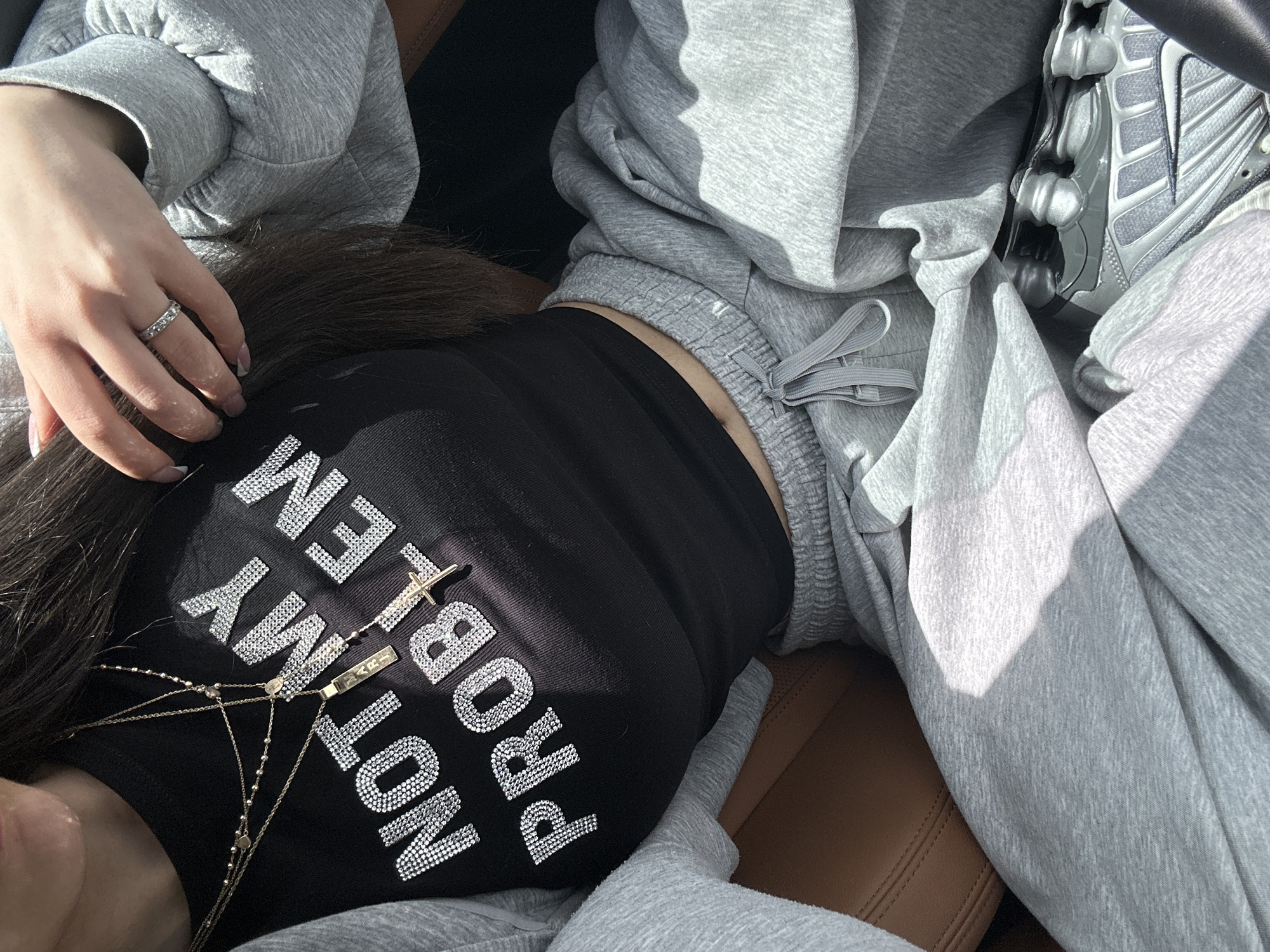 The image shows a person sitting in what appears to be a car seat, likely made of tan leather. They are wearing a gray hoodie and sweatpants. A black tank top with the phrase "NOT MY PROBLEM" written in silver rhinestones is visible. The person has dark hair and is wearing a silver ring on their finger. A silver Nike shoe is also visible. The location cannot be determined based on the information provided.