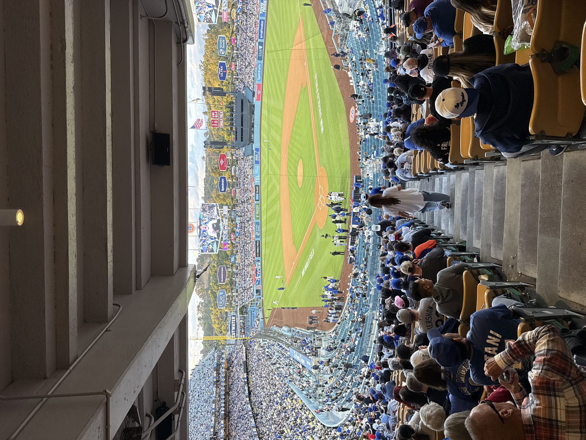 This is an image taken from a high-angle perspective inside a baseball stadium, filled with a dense crowd of spectators. The image shows a baseball field where a game is being played.
The spectators are in stadium seats and are wearing a variety of clothing. Some fans are wearing baseball caps and jerseys, likely in support of the home team. The field itself is green with dirt base paths. The outfield walls are adorned with advertising banners. In the background beyond the outfield, there are buildings and greenery, indicating an urban setting. The image seems to capture a lively and densely populated baseball game.
This location is in the city of Los Angeles, United States of America.