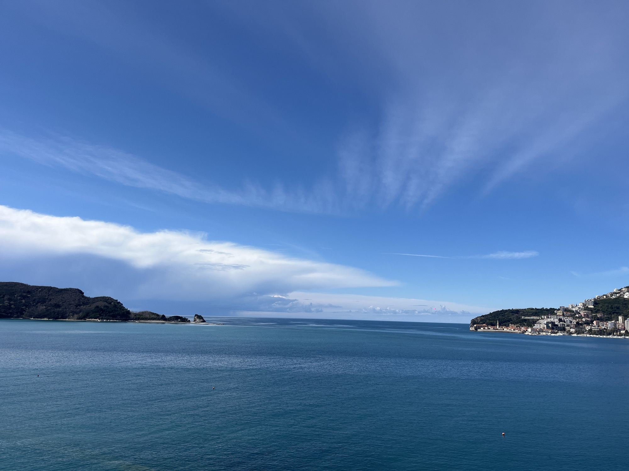 The image presents a wide panoramic view of the Adriatic Sea and the coastline of Budva, Montenegro. The scene is dominated by a clear, bright blue sky containing scattered, wispy cirrus clouds, with a larger band of cumuliform clouds visible on the distant horizon to the left. The water, a deep teal-blue, occupies the majority of the foreground and midground, exhibiting minimal surface disturbance.
On the far left, a dark, undulating landmass, heavily vegetated with trees, extends into the sea, indicative of Sveti Nikola Island or a similar peninsula. Small, rugged rock formations are visible off its eastern tip. On the far right, the historic Old Town of Budva is situated on a prominent peninsula, characterized by a dense cluster of light-colored buildings with distinct reddish-brown tiled roofs. The cityscape extends eastward up a green, forested hillside, featuring numerous modern multi-story buildings integrated into the natural landscape. At the base of the Old Town peninsula, a marina containing multiple white, docked recreational boats is discernible. Two buoys are present in the water: one dark, circular buoy in the left midground and one smaller, reddish-orange spherical buoy in the right foreground. No individuals, animals, or active watercraft are visible within the frame.