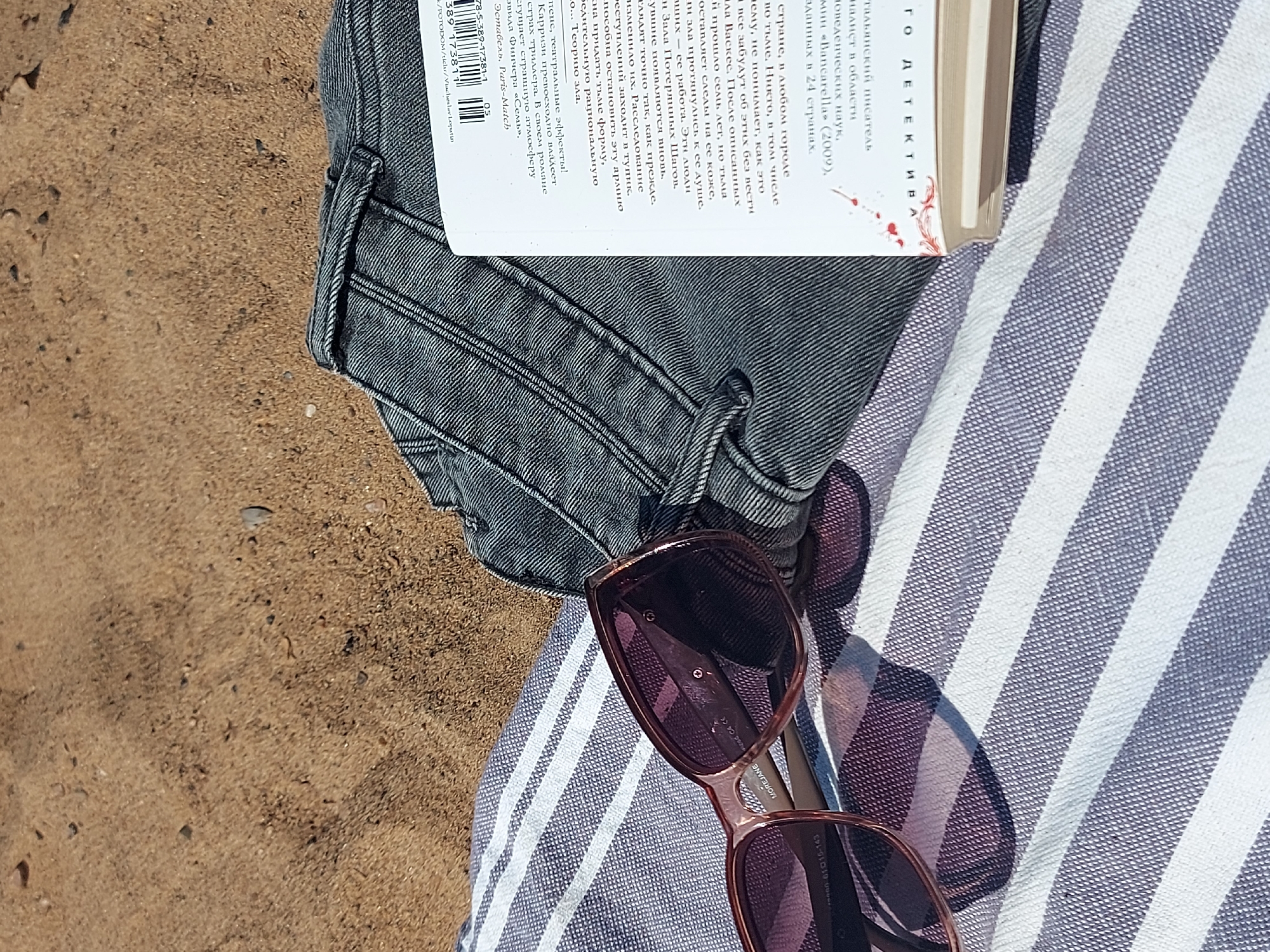 This image appears to be taken on a beach. The scene includes several objects: a book in Russian (likely a detective novel), a pair of sunglasses, and a pair of gray denim shorts, all resting on a striped towel. The environment consists of sand, suggesting a beach setting.