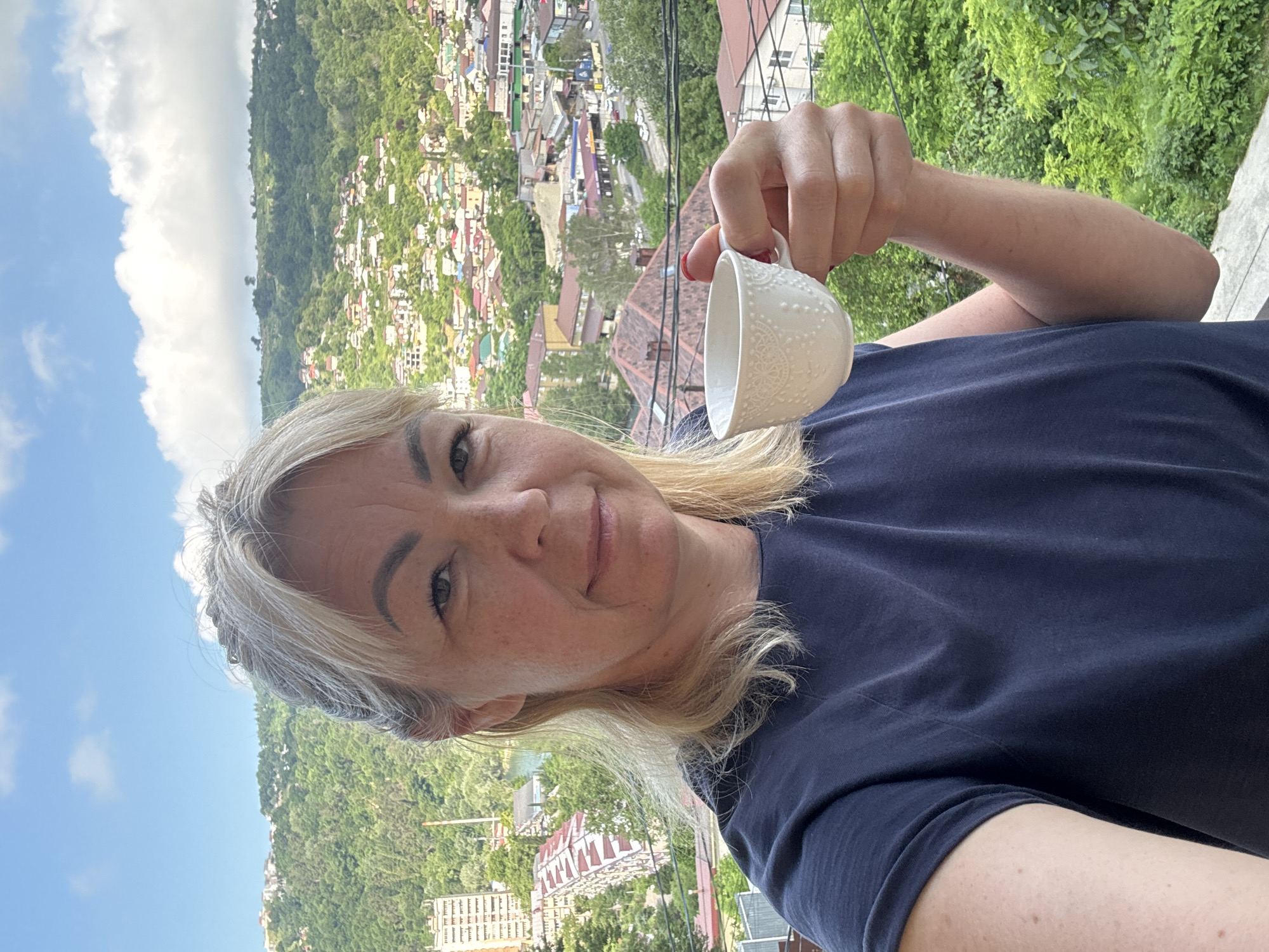 A blonde woman with light skin and freckles takes a selfie while holding a white teacup with an embossed pattern. She is wearing a dark blue t-shirt. Her hair is pulled back, with some strands loose around her face. She is looking at the camera with a slight smile. The background is a cityscape with buildings and lush green hills, characteristic of Sochi, Russia. The sky is blue with scattered white clouds. The woman is positioned in the foreground, with the city and natural landscape behind her.