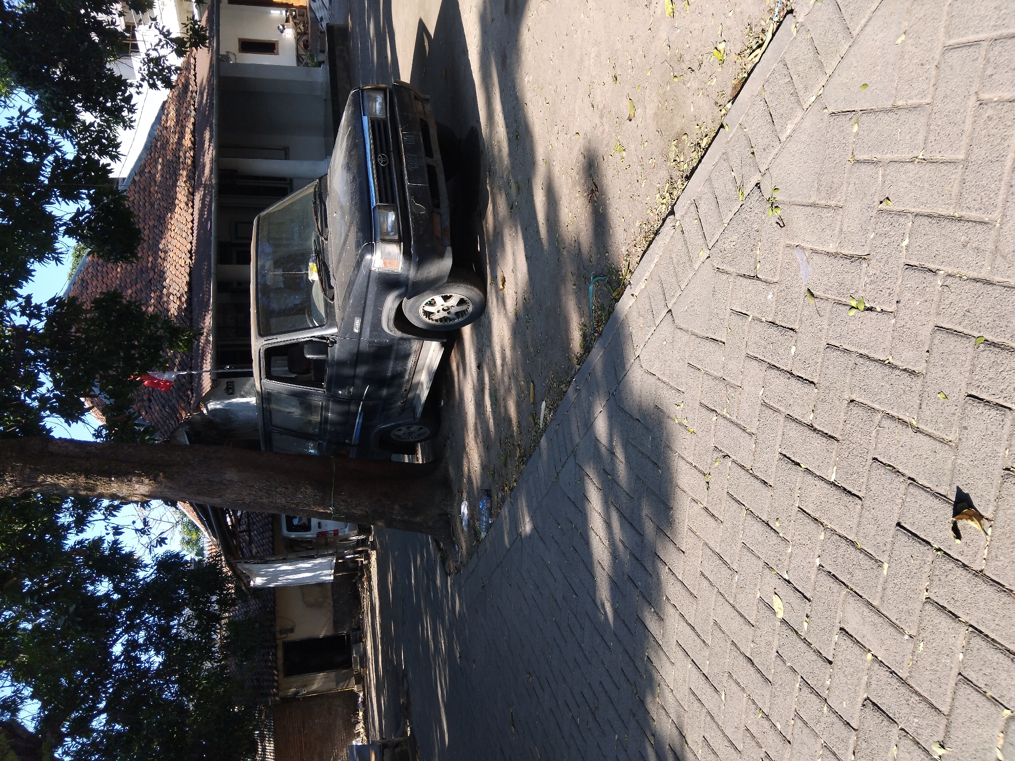 An outdoor, eye-level, medium shot shows a black four-door SUV parked on a dirt lot next to a paved walkway in Kepuh Kiriman, Indonesia. The SUV is angled away from the viewer and partially obscured by a large tree with dark green leaves. The vehicle has a dark-colored grill and headlights, and its wheels are visible. In the background, a single-story building with a red tile roof is visible, along with a brick wall and another smaller white building. The ground in the foreground is covered with grey interlocking pavers, and the area around the car is dirt with scattered leaves and debris. Sunlight casts shadows from the tree and leaves onto the ground.