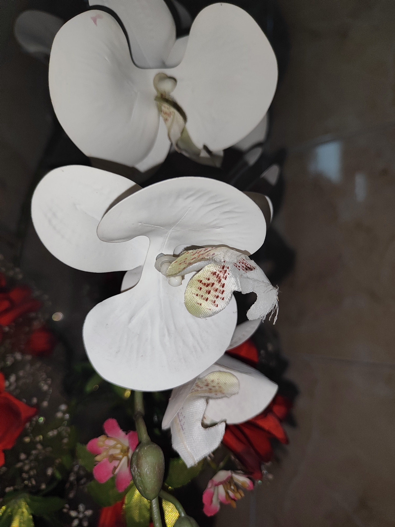 This is a close-up, vertical, eye-level, indoor, artificial lighting, low-angle shot of several artificial white orchid flowers. The orchids are arranged in a bouquet with other artificial flowers, including red and pink ones, and some small white buds. The background is a blurred, mottled brown surface, possibly a wall or furniture. The image appears to have been taken in Bondowoso, Indonesia, though no specific details about the location within the city are discernible.