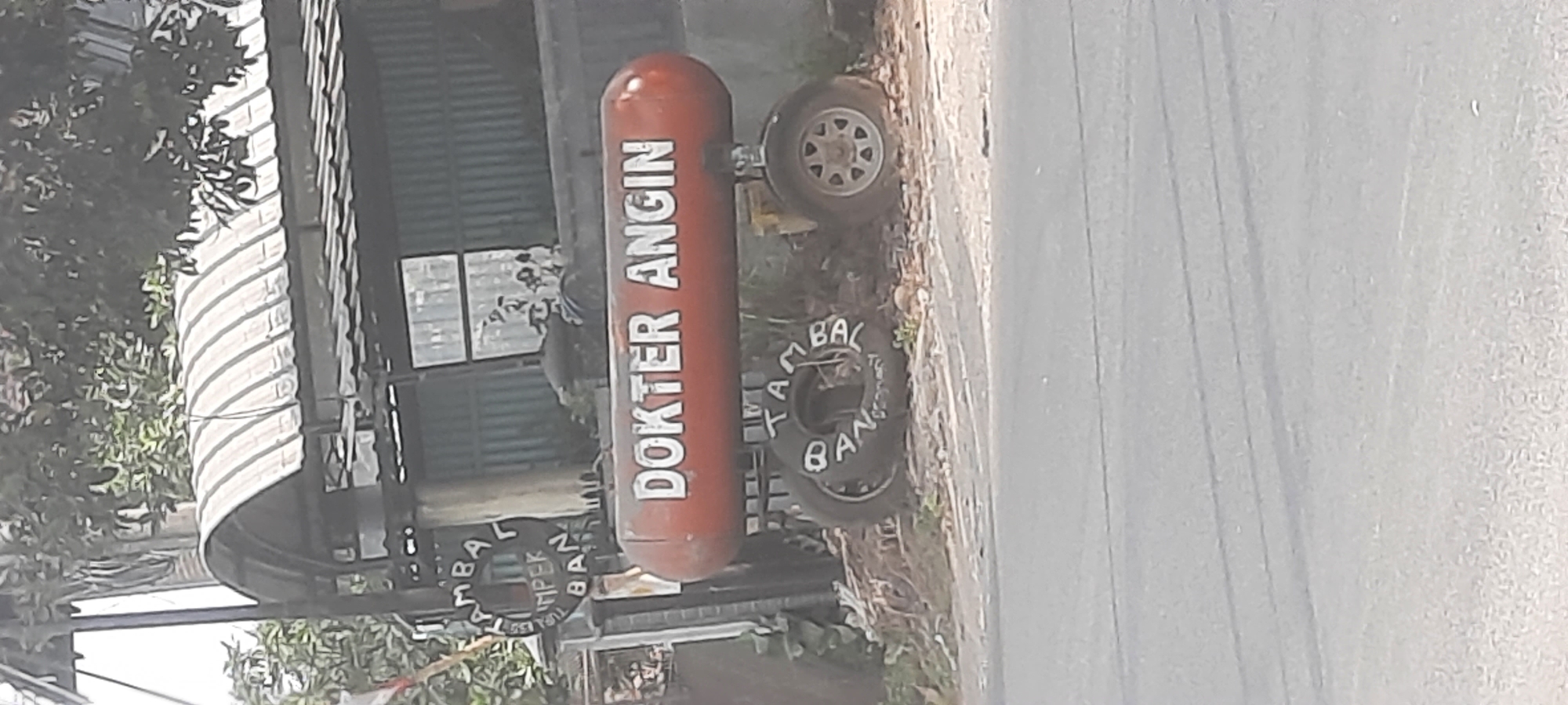 In Siraman, Indonesia, a red cylindrical object, likely a repurposed air tank, is mounted on a wheeled chassis and serves as a sign. White text on the tank reads "DOKTER ANGIN," which translates to "Wind Doctor" or "Air Doctor." The object is positioned in front of a structure with a corrugated metal roof and a green corrugated metal wall. In the foreground, a paved road is visible. The ground around the tank is covered with debris and vegetation. On the left side of the tank, a circular sign with text that appears to be "BAMBA" and "IMPER" is affixed to a pole. Tires with white lettering are also visible on the chassis, with one tire displaying text that reads "BAN TA BAL." The overall scene suggests an outdoor setting, possibly a roadside business or workshop.