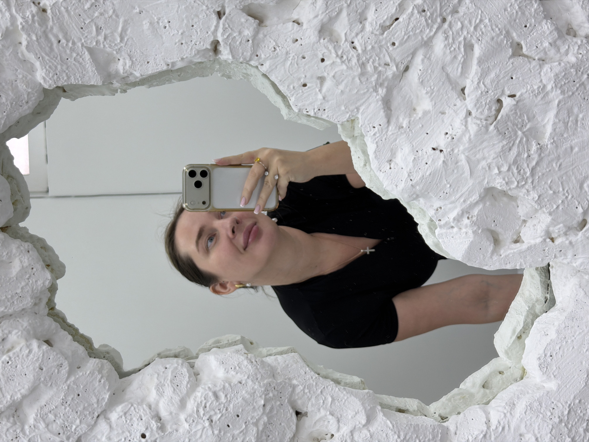 A woman in a black top is reflected in a white, irregularly shaped mirror with a textured frame. She is holding a mobile phone, presumably to take a photograph. The reflection also shows a white wall in the background. There is no information to verify this is in the city of Moscow, Russia.