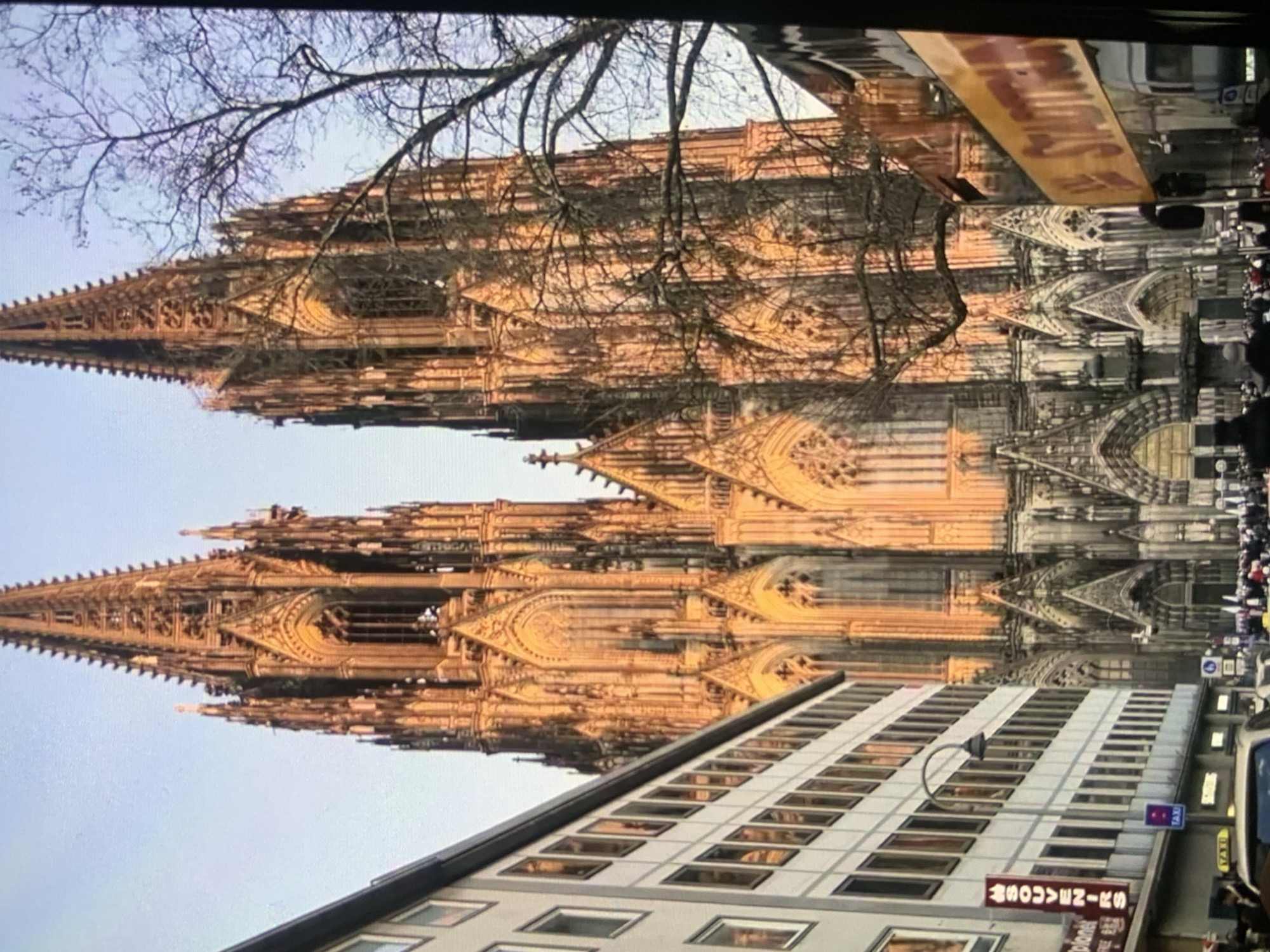 The image shows the exterior of a large, gothic cathedral with two tall spires. The cathedral is made of stone and is ornately decorated with intricate carvings and architectural details. Sunlight illuminates the facade, casting a warm glow. In the foreground, there is a street scene with buildings, people, and vehicles. A multi-story building with many windows is visible on the left, and a bus or large vehicle with a yellow sign is on the right. A crowd of people is gathered on the street in front of the cathedral. The sky above is clear and light blue. The image appears to be taken in a European city given the architectural style of the cathedral and the presence of a "SOUVENIRS" sign. The location is not Stavropol, Russia.