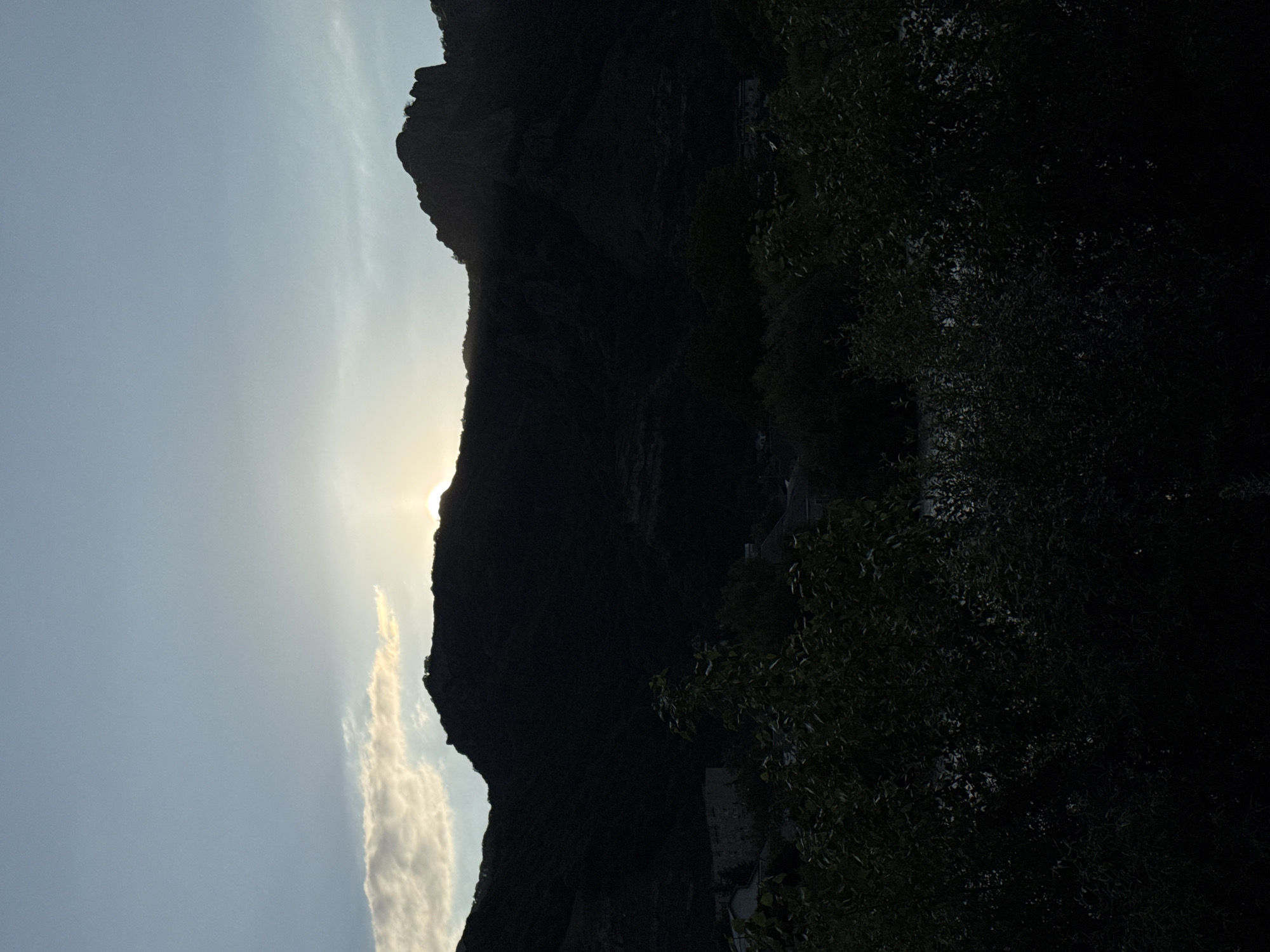 This is a silhouette image of a mountainous landscape at dusk or dawn. The sun is low on the horizon, peeking over the crest of the mountains, casting them in shadow. A single, wispy cloud is illuminated by the sun. In the foreground, there is dense foliage, obscuring the ground and any potential inhabitants or structures. The sky transitions from a pale blue at the top to a darker hue closer to the horizon. Based on the visual cues of the terrain and vegetation, it is plausible this scene is located in or around Karachayevsk, Russia, a city known for its mountainous surroundings.