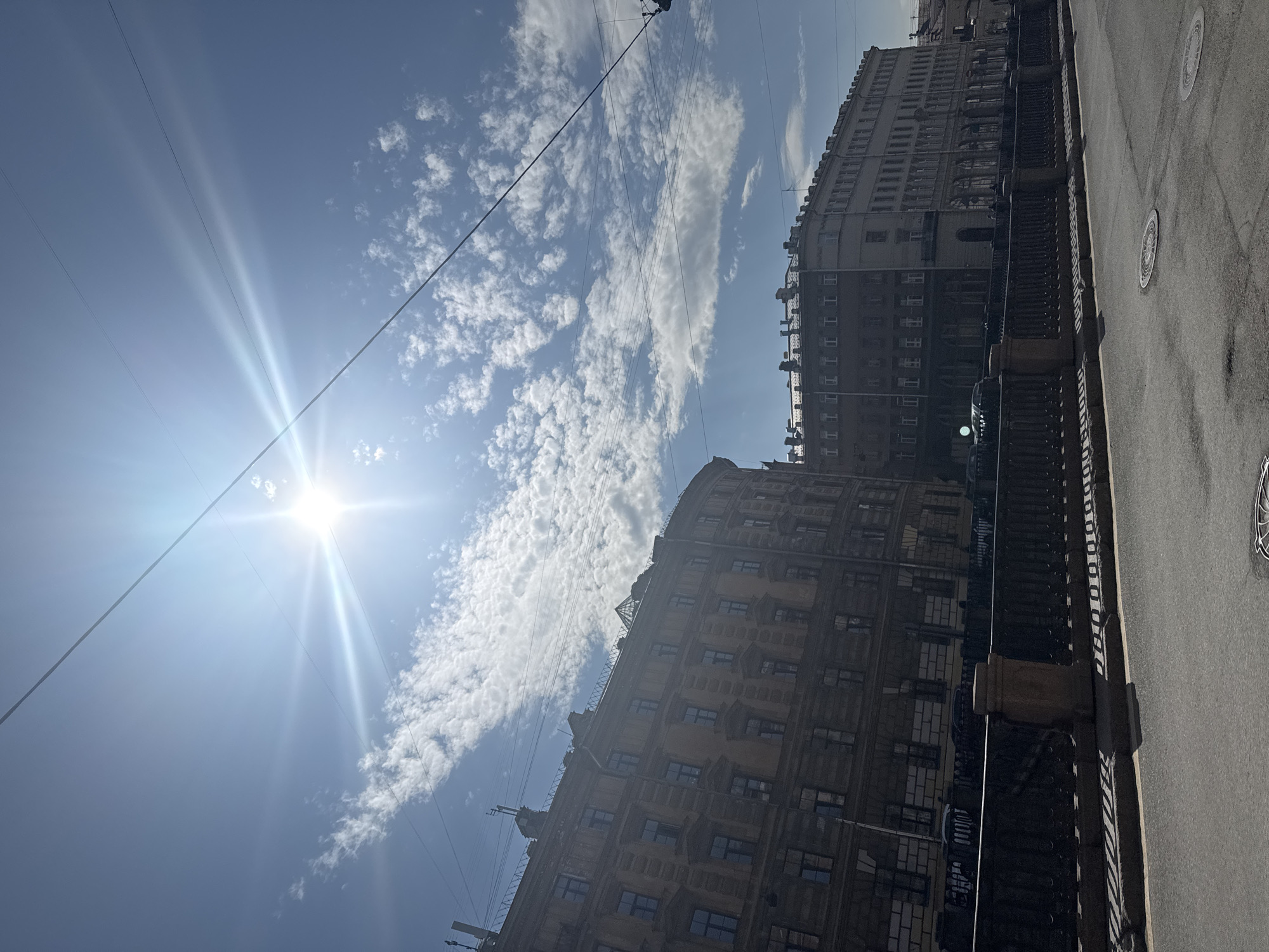 This image captures a vibrant urban street scene, oriented to show the sky, sun, and prominent architecture. The perspective is upward-angled, emphasizing the grandeur of the buildings and the expanse of the sky.
The upper portion of the image is dominated by a bright blue sky, adorned with scattered, elongated patches of white cumulus clouds. A brilliant sun is positioned in the upper right, casting intense rays of light and producing a distinct lens flare. Several thin, dark utility wires crisscross the sky, adding an element of urban infrastructure.
On the right side, a large, multi-story building stands prominently. Its facade appears to be made of light-colored stone or stucco, possibly yellowish or beige, and features numerous rectangular windows, many with intricate frames. The architectural style suggests a historical period, characterized by detailed cornices and a grand scale. Portions of other buildings are visible in the background, further hinting at a dense urban environment.
At the bottom of the frame, a wide, grey paved surface, likely a street or sidewalk, extends into the distance. A dark, ornate metal railing or fence runs parallel to the buildings, separating the paved area from the structures. Two circular manhole covers with geometric patterns are visible on the pavement.
No specific activity or event is taking place; the scene is static. The clear blue sky and bright sun suggest a pleasant daytime, likely mid-morning or afternoon. The architectural style of the buildings, combined with the presence of ornate railings and a wide thoroughfare, strongly aligns with the provided location context of Saint Petersburg, Russia. There is no visible text in the image.