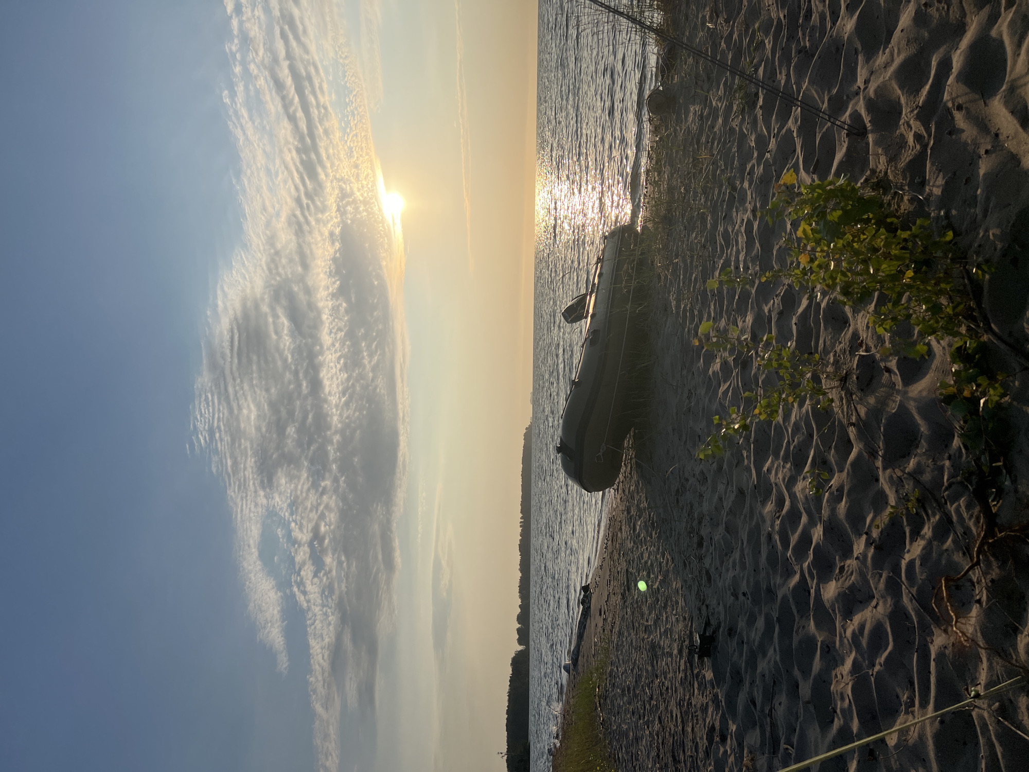 This image depicts a tranquil scene on a sandy beach with an inflatable boat resting on the shore. The sun is setting, casting a warm glow across the water and sky, with the sun partially obscured by wispy clouds. The sea is calm, with gentle waves lapping at the shore. In the background, a treeline is visible on the horizon, suggesting a natural landscape. The sand is textured with ripples, indicating the presence of wind. A fishing rod can be seen planted in the sand near the boat. The overall atmosphere is serene and peaceful, evoking a sense of quiet contemplation. The location is identified as the city of Borovoe, Russia.