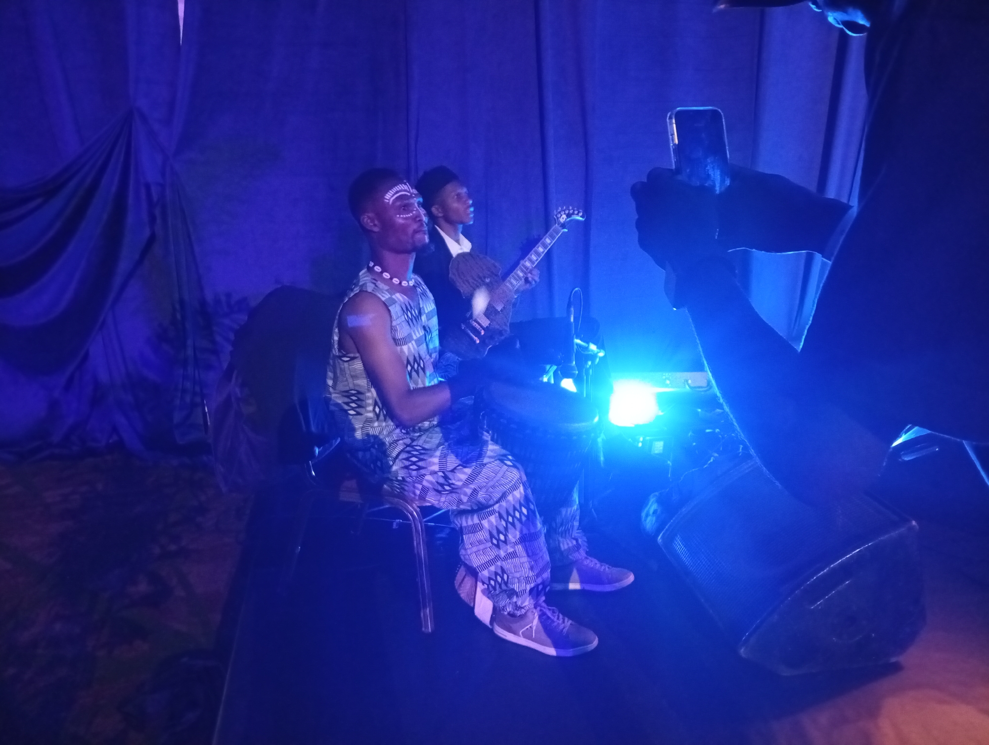 In a blue-lit indoor setting in Itirin, Nigeria, a person dressed in traditional clothing with face paint sits on a chair with a drum between their legs. A second individual stands behind them playing an electric guitar. A third person stands to the right, holding up a smartphone as if taking a photo or video of the scene. A speaker is on the floor in front of the individuals. A dark fabric drape is visible in the background.