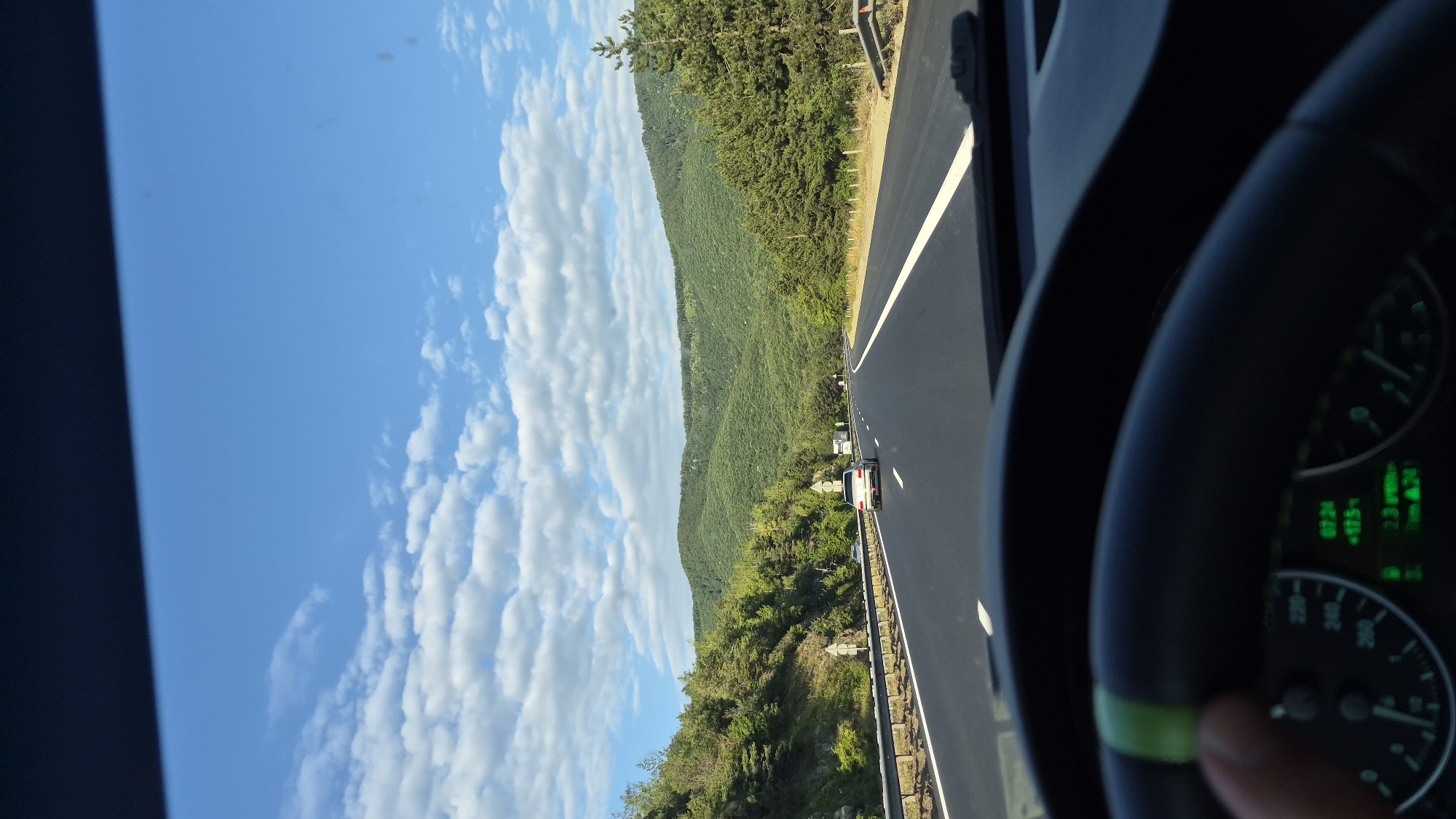 The image shows a view from the driver's seat of a car traveling on a highway. The highway curves to the right and is lined with guardrails and signs. Green, rolling hills covered in trees form the backdrop. The sky is blue with scattered white clouds. The dashboard, including the steering wheel and instrument cluster, is visible in the foreground. The instrument cluster shows a speed of approximately 125 and the time 07:24. A hand with a green stripe on the steering wheel is partially visible. Multiple cars are on the highway ahead. The scene is a scenic, rural highway on a clear day.