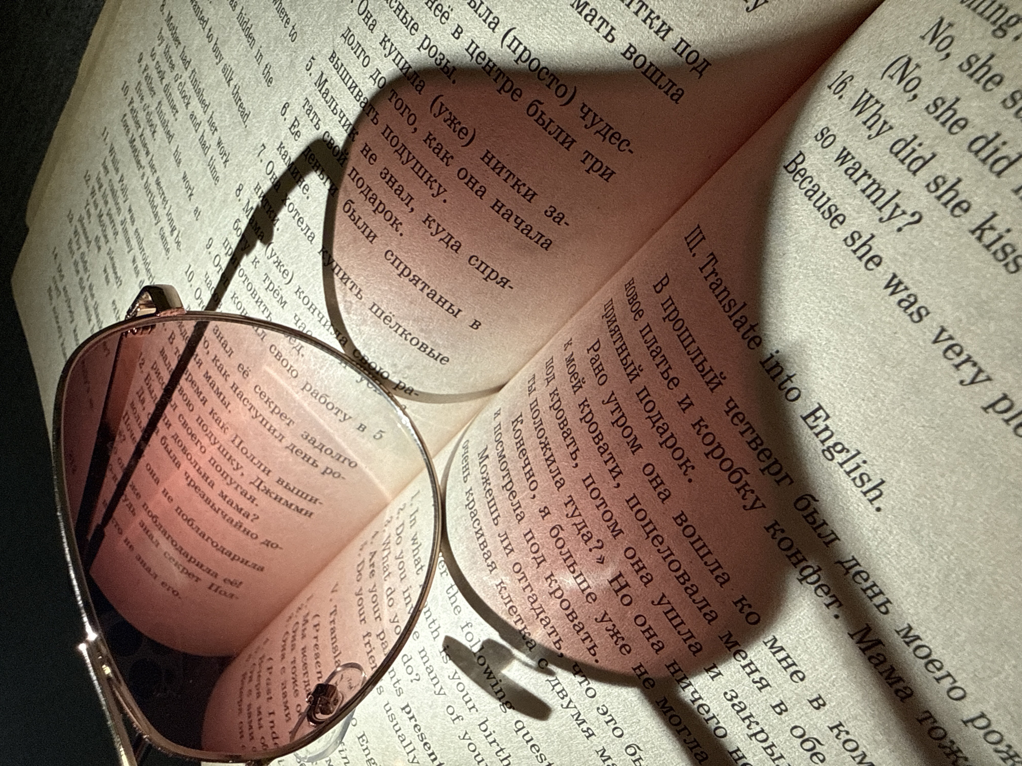 An open book lies on a dark surface, with a pair of sunglasses resting on its pages. The book appears to be a language learning text, containing both Russian and English text. The lighting creates strong shadows on the pages. The environment is a close-up, indoors setting, with nothing discernible to indicate the image was taken in Dmitrov, Russia.
