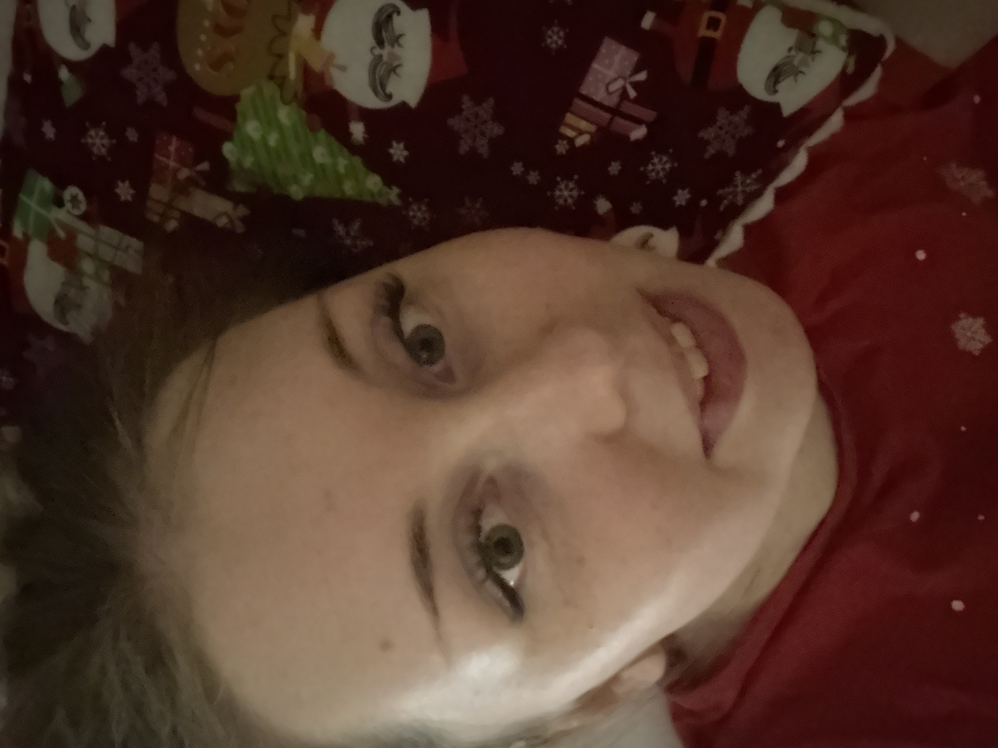 A close-up, rotated view depicts a person lying down. Their face, with visible freckles and eye makeup, is prominently featured. The person is wearing a red shirt with white snowflake patterns. Beside them is a pillow with a Christmas-themed pattern including Santa Claus, snowflakes, and gift boxes. The location and city are unknown.