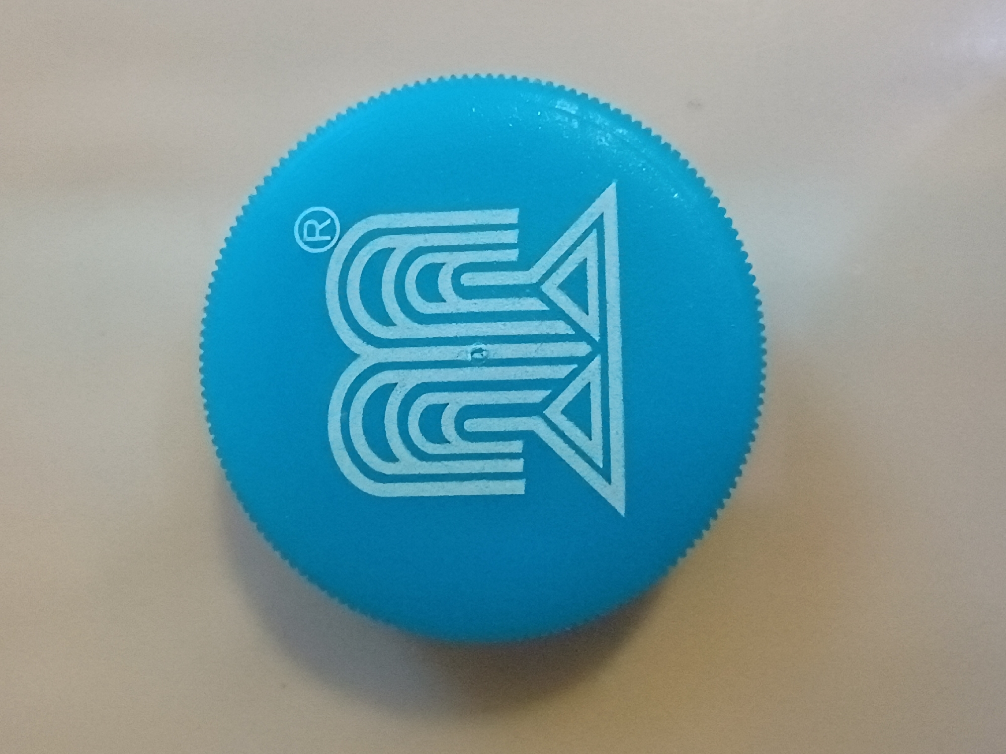 The image displays a close-up, top-down view of a bright blue, circular plastic cap. The cap has a serrated edge, suggesting it's a lid for a container. Prominently featured on the cap is a white, stylized logo. The logo consists of concentric, curved lines forming a shape reminiscent of the letter "B" or an abstract, upward-pointing arrow with internal divisions. To the left of the main logo, a smaller registered trademark symbol "®" is visible. The cap rests on a light-colored, textured surface, possibly a table or countertop. The lighting suggests an indoor setting, likely during the daytime. No people are visible in the image. The context of Zaporizhzhia, Ukraine, does not provide specific visual cues within the image itself, but it sets a geographical location for where this object might be found. There is no discernible weather or time of day information from the visual elements alone. The image is static, showing a single object without any action.