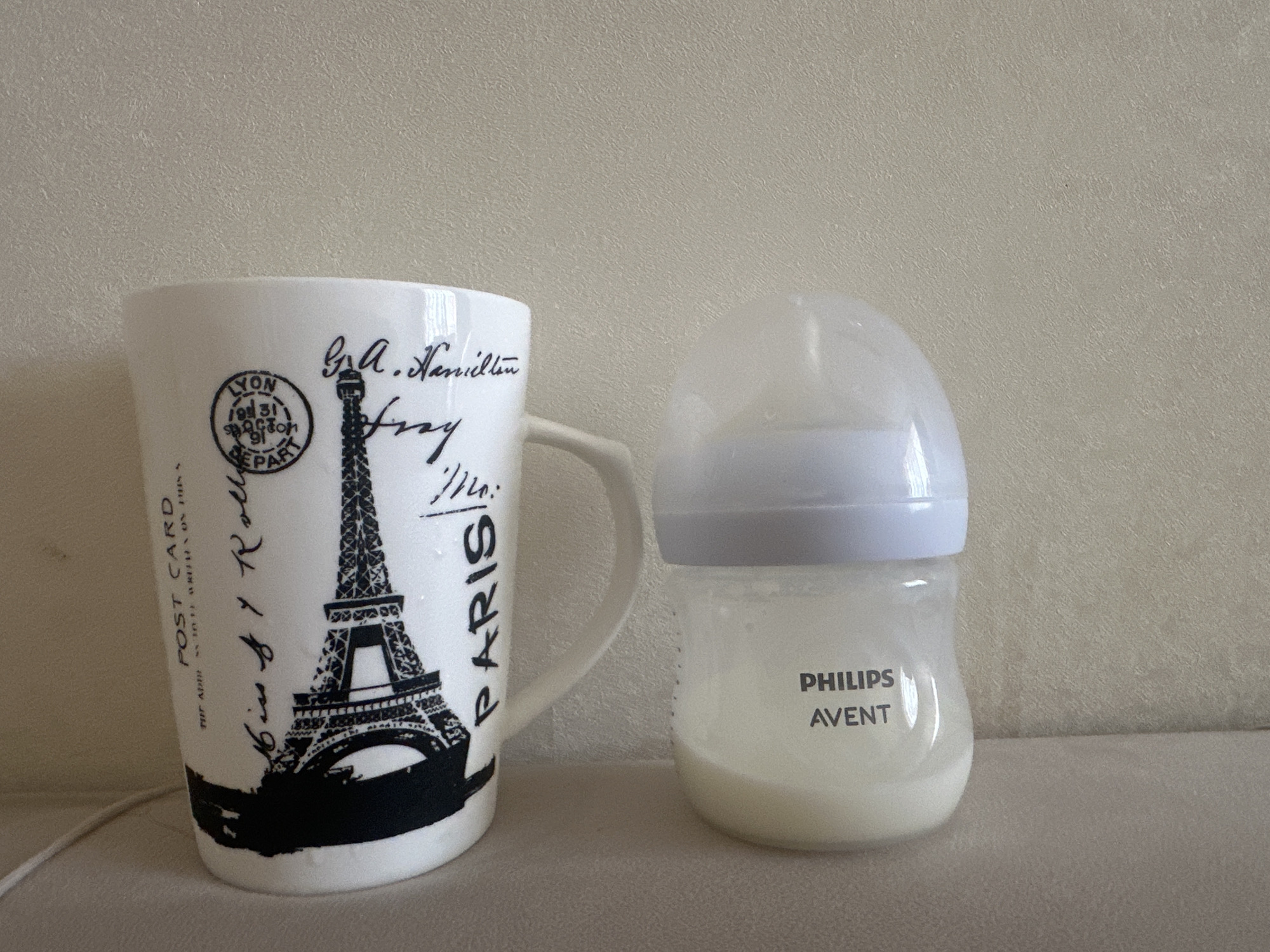 A white ceramic mug with a black Eiffel Tower design and French text, and a clear plastic baby bottle with a white cap containing a pale yellow liquid are placed side-by-side on a beige textured surface. The mug is on the left, and the baby bottle is on the right. The background is a textured beige wall. There are no people in the image. The image does not provide any information or context that would suggest a location in Masazir, Azerbaijan.