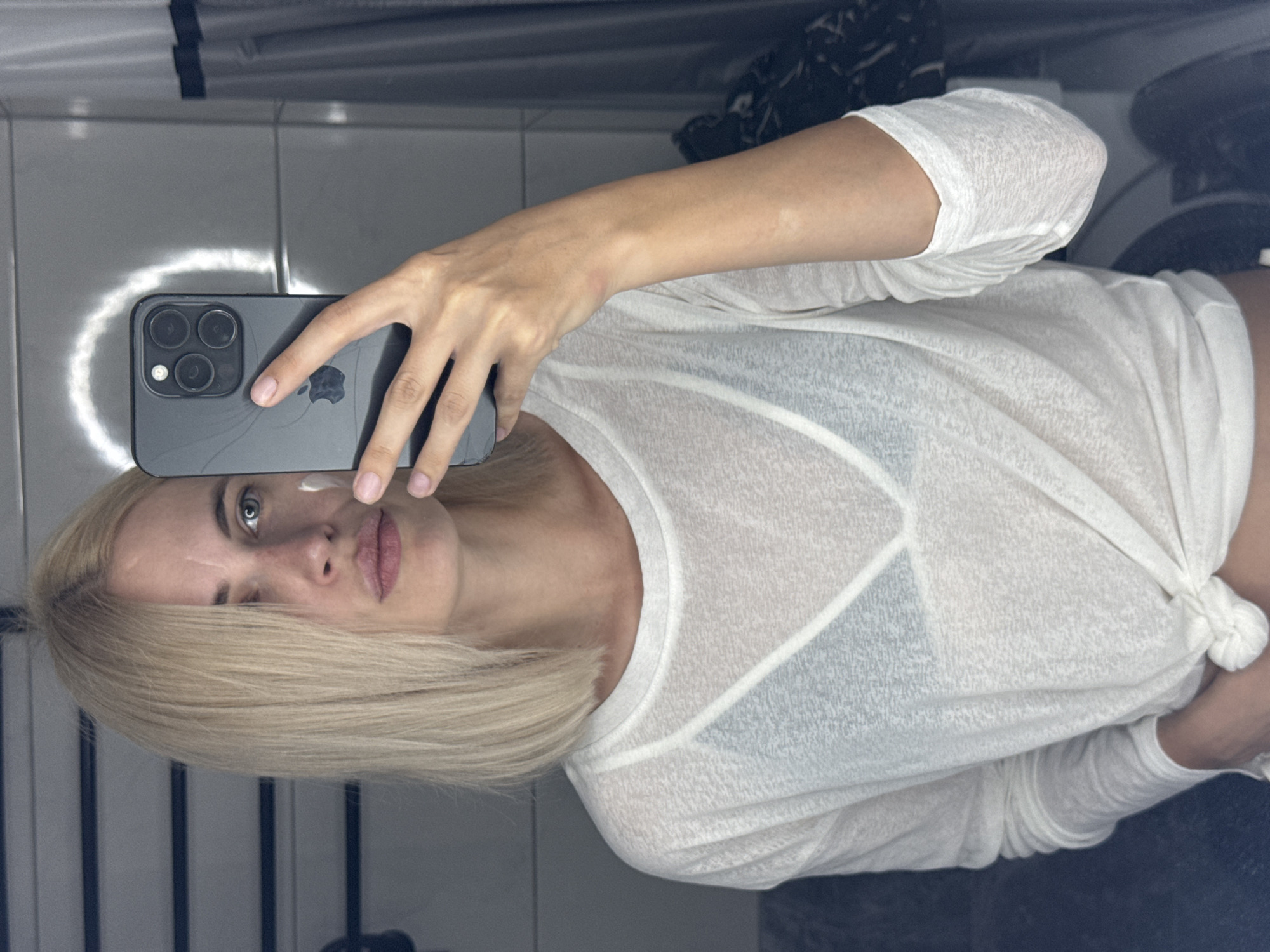 A woman with blonde, shoulder-length hair is taking a selfie in a bathroom. She is wearing a white, sheer, long-sleeved shirt tied at the waist over a dark-colored bra. A small dab of white cream is visible on her cheek. She is holding a smartphone in her right hand, angled to capture her reflection. The background shows tiled walls and a shower curtain. The image does not contain enough information to determine the specific location within Saratov, Russia.
