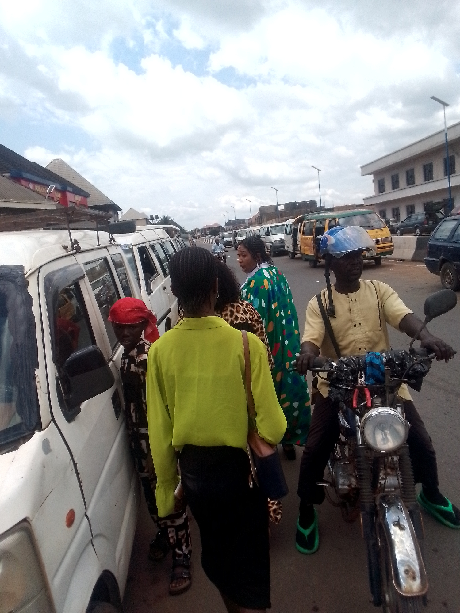 The image depicts a street scene in an urban location, likely in Unknown, Unknown. A row of white vans lines the left side of the street, with some of them appearing to be public transportation vehicles. A yellow and green public transport bus is visible in the background.
Several people are in the foreground. A person in a green shirt and black pants stands facing away from the camera. Next to them is a person wearing a red head covering and patterned shirt standing next to one of the white vans. Another person in a green and blue patterned dress is standing nearby. A man wearing a yellow shirt, blue helmet, and green sandals is seated on a motorcycle. He is holding the handlebars. There is a pair of pliers attached to the front of his motorcycle.
The environment includes buildings in the background, one of which is a light-colored structure with multiple windows. Streetlights are also visible in the distance. The sky is overcast with cloudy conditions.