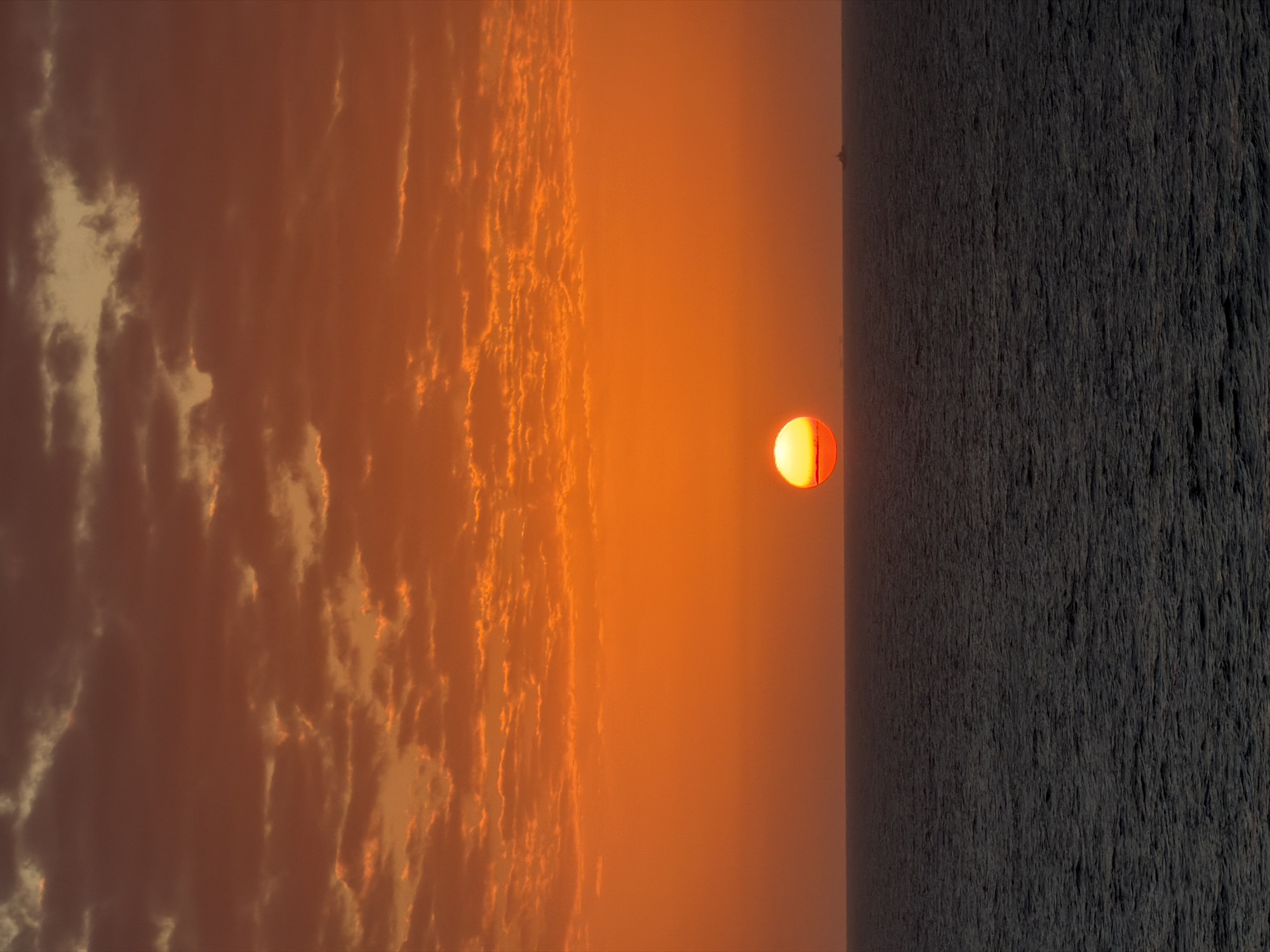 The image depicts a sunset over an ocean. The sun is positioned in the center of the frame, appearing as a semi-circular orange and red disc. The sky on the left side of the sun is filled with textured clouds, bathed in warm orange and red hues. To the right of the sun, the ocean stretches out, appearing dark and textured, with the waves casting shadows. The location and city are unknown.