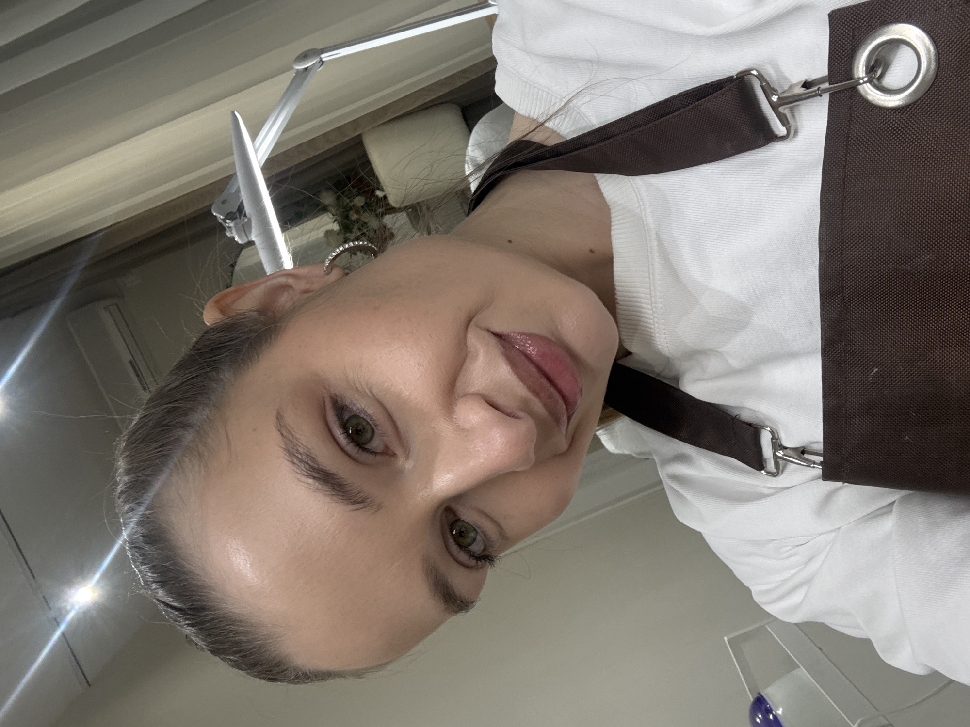 A close-up, canted, indoor shot shows a fair-skinned woman with green eyes and light brown hair pulled back. She is wearing a white shirt and a brown apron, and has a small, clear hoop earring. In the background, out of focus, are a window with a plant and a lamp. There is no indication of the city of Budva, Montenegro in the image.