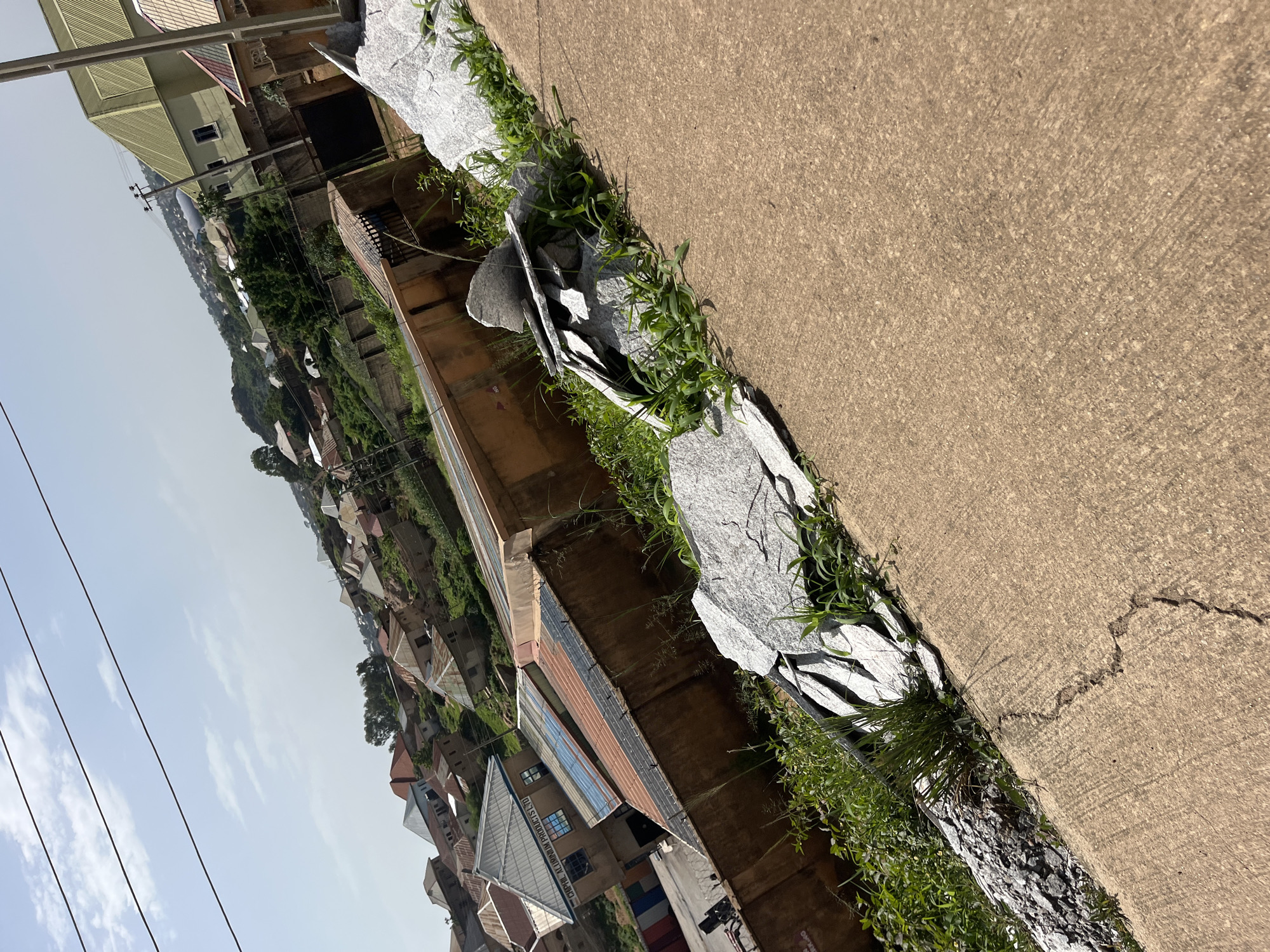 The image depicts an upward view of a residential area with a collection of buildings, suggesting a dense urban or semi-urban environment. The structures vary in architectural style, with some featuring corrugated metal roofs in shades of red, blue, and grey, while others have more modern designs with light-colored roofs. The buildings are situated on a hillside, creating a tiered effect, and are interspersed with greenery, indicating a possibly uneven terrain. In the foreground, there is a concrete pathway or road, with some scattered pieces of light-colored stone or concrete debris and overgrown grass. The sky is visible in the upper portion of the image, with scattered clouds. The overall scene conveys a sense of everyday life in a developing urban landscape. The image was taken in Gura Top, Nigeria.