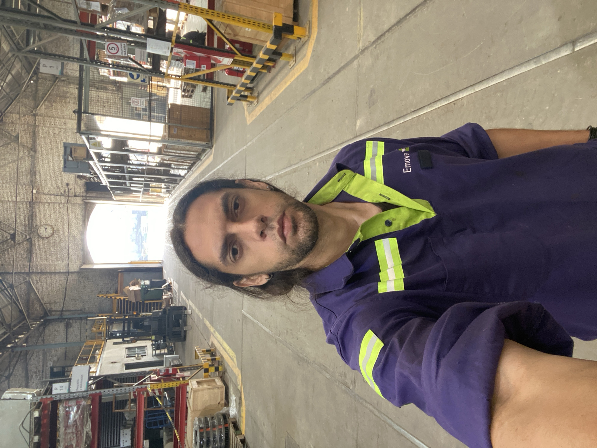 A man is standing in a large, industrial warehouse-like building in Sáenz Peña, Argentina. The building has a high ceiling with exposed brick and metal support beams. There is natural light coming from a large opening at the far end of the building. The man is wearing a purple and lime-green work uniform with the word "Emova" on the chest. He is looking at the camera. The floor is concrete with yellow lane markings. Shelving units filled with various items are visible along the sides of the building. There are safety barriers constructed of yellow and black metal tubing. A clock is visible on the upper wall near the back of the warehouse.