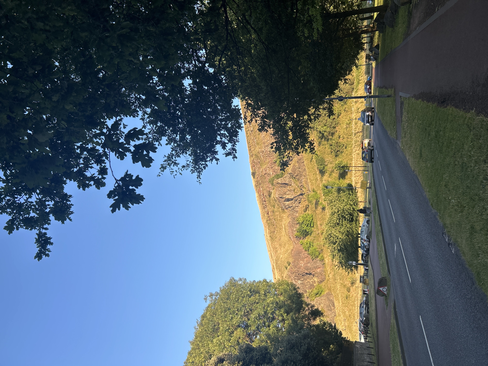 The image depicts a roadside scene in the City of Edinburgh, United Kingdom, likely near Arthur's Seat, given the prominent rocky hill in the background. On the left side of the frame, a paved road runs parallel to a green grassy verge. Several cars are parked on the opposite side of the road in a designated parking area. Beyond the parking lot, a large, tree-covered hill rises, its slopes illuminated by the warm light of what appears to be late afternoon or early morning sun, casting shadows and highlighting the rocky outcrops. To the right of the road, a red-surfaced pedestrian walkway is visible, bordered by grass and large rocks. Mature trees with dark green foliage line the right side of the walkway, their branches extending towards the clear blue sky. One individual, dressed in dark clothing, is walking away from the camera on the pedestrian path. The overall impression is one of a tranquil urban park setting adjacent to a natural landscape.
