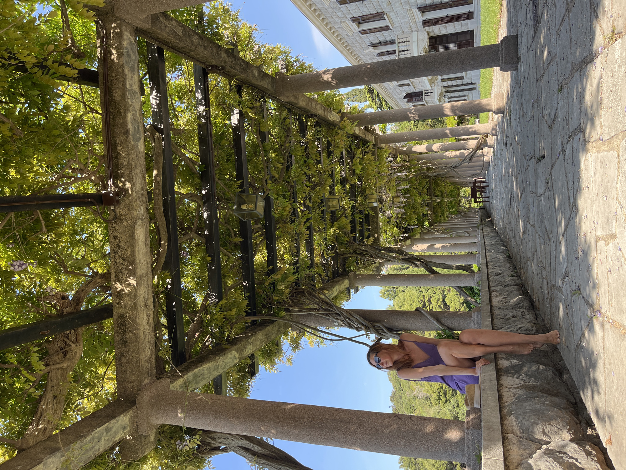 This image captures a sunny outdoor scene, likely within the scenic grounds of Sveti Stefan, Montenegro. A woman, wearing a purple tank top or dress and sunglasses, is seated casually on a low stone ledge under a pergola. She is barefoot and her legs are bent, facing slightly away from the camera.
The pergola, constructed from robust stone columns and overhead beams, is heavily adorned with vibrant green foliage, possibly wisteria, which drapes across the structure. Clusters of purple flowers are interspersed among the leaves. Bright sunlight filters through the canopy, creating a play of dappled shadows on the stone-paved walkway beneath.
In the background, a grand, light-colored building with intricate architectural details, multiple windows, and dark shutters stands prominently under a clear blue sky. Distant green hills or lush trees are visible beyond the structures. The scene conveys a sense of leisure and tranquility in a beautiful, historic setting. No discernible text is visible in the image.