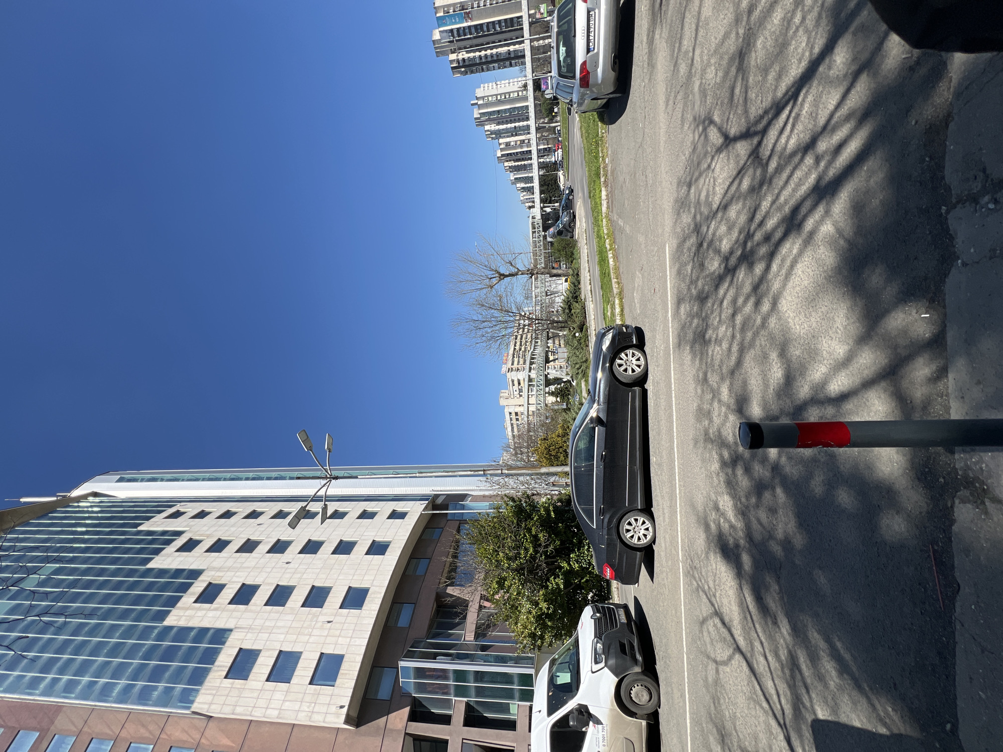 The image depicts a street scene in Burgas, Bulgaria. A black car is parked next to a white van on a gray asphalt street. Adjacent to the buildings are large office buildings with many windows, and across the road are apartment buildings. A tall metal light pole stands near the office buildings. Tree shadows are cast on the street. A gray pole with a red stripe is on the right edge of the frame. The sky is clear and blue. There are no people visible.