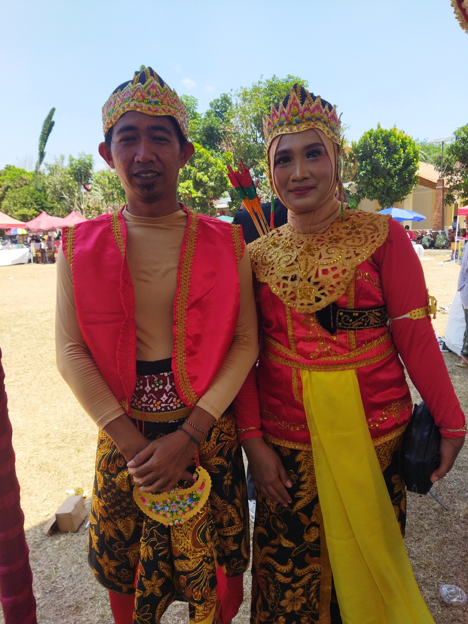 Two individuals, a man and a woman, are posing for a photograph outdoors. They are dressed in traditional, ornate clothing. The man wears a tan long-sleeved shirt, a bright red vest adorned with gold trim, and a patterned kilt-like garment with black and gold floral motifs. He also wears a decorative crown. The woman is wearing a tan headscarf, a red long-sleeved top with intricate gold embroidery around the neckline and sleeves, a yellow sash, and a matching patterned kilt-like garment. She also wears a crown and a gold necklace-like decoration. They are standing in an open, grassy area, with trees and some tents visible in the background. The sky is clear and blue. The overall impression is that they are participating in a cultural event or festival. The location is identified as Semarang, Indonesia.