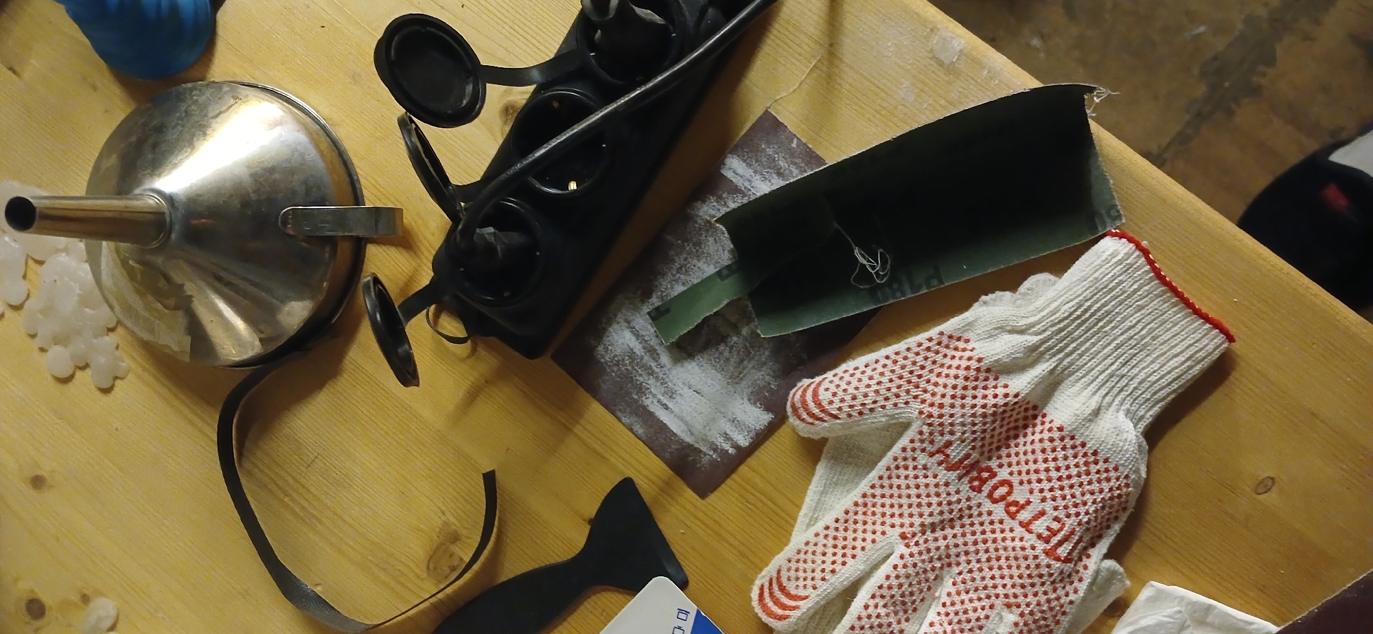 The image presents an overhead view of a wooden workbench or table laden with various tools and materials, suggesting a crafting, DIY, or repair project.
In the upper-left, a shiny silver metal funnel with a narrow spout and a small handle is visible. Below it, several small, white, translucent, irregularly shaped objects, possibly silicone molds or cast items, are scattered. A blue object is partially seen at the top-left edge.
Towards the center, a black multi-socket power strip with several circular, flip-open covers is present, alongside a black cable. To its right, two pieces of sandpaper are laid out: one dark green with "P180" partially visible, and another darker piece covered in white sanding dust, indicating recent use. A tangled white string rests on the green sandpaper.
In the bottom-right, a white fabric work glove with a ribbed cuff and numerous red rubber dots on the palm and fingers is displayed. The Cyrillic text "ПЕТРОЯР" (PETROYAR) is clearly printed in red on the palm, which aligns with the provided location context of Moscow, Russia.
A black, flat, spatula-like scraper tool is in the bottom-center, partially obscuring a white card with faint text like "DD" and "10". A thin black strip of material, possibly electrical tape, extends across the lower-left. A crumpled white tissue is visible at the very bottom-right.
The lighting is artificial and warm, typical of an indoor setting, possibly an evening or night work session. No individuals are visible, and no specific emotions are conveyed. The arrangement of items points towards an activity involving sanding, pouring, and possibly electronics or molding.