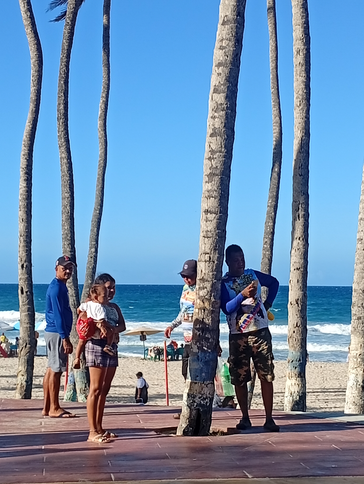 The image shows a sunny day at Playa El Agua, Venezuela. Five adults and one child are visible on a raised walkway. Several tall, slender palm trees are present between the foreground and the beach. The sea is in the background with gentle waves breaking on the shore. Beach umbrellas and chairs are visible on the sand. The adults are engaged in conversation. One woman is holding a small child. The adults' clothing ranges from casual shorts and t-shirts to long-sleeved shirts and caps.