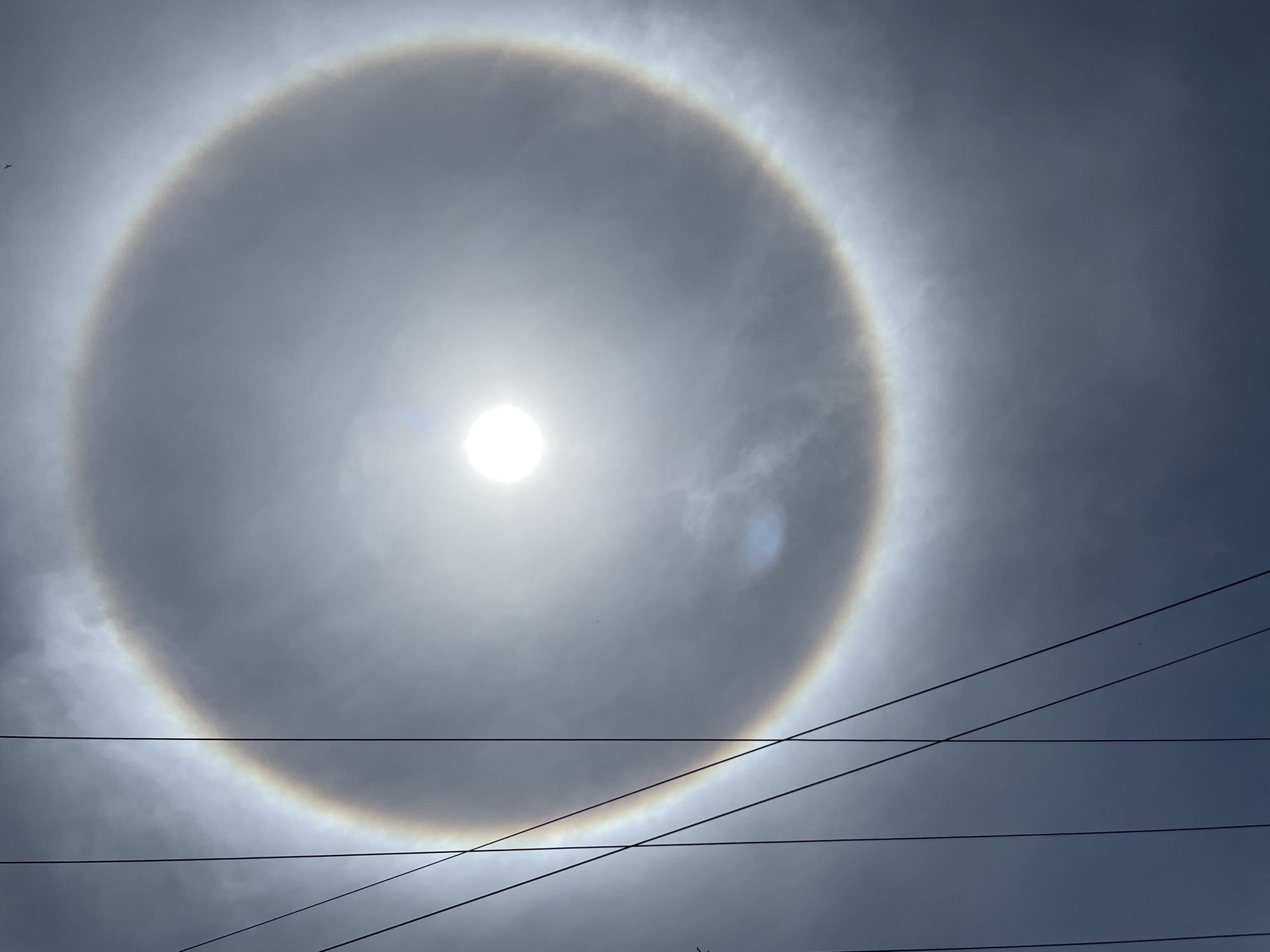 A circular halo surrounds the sun in a cloudy sky. Power lines stretch across the lower portion of the image. The location, Porlamar, Venezuela, is not discernible from the image itself.