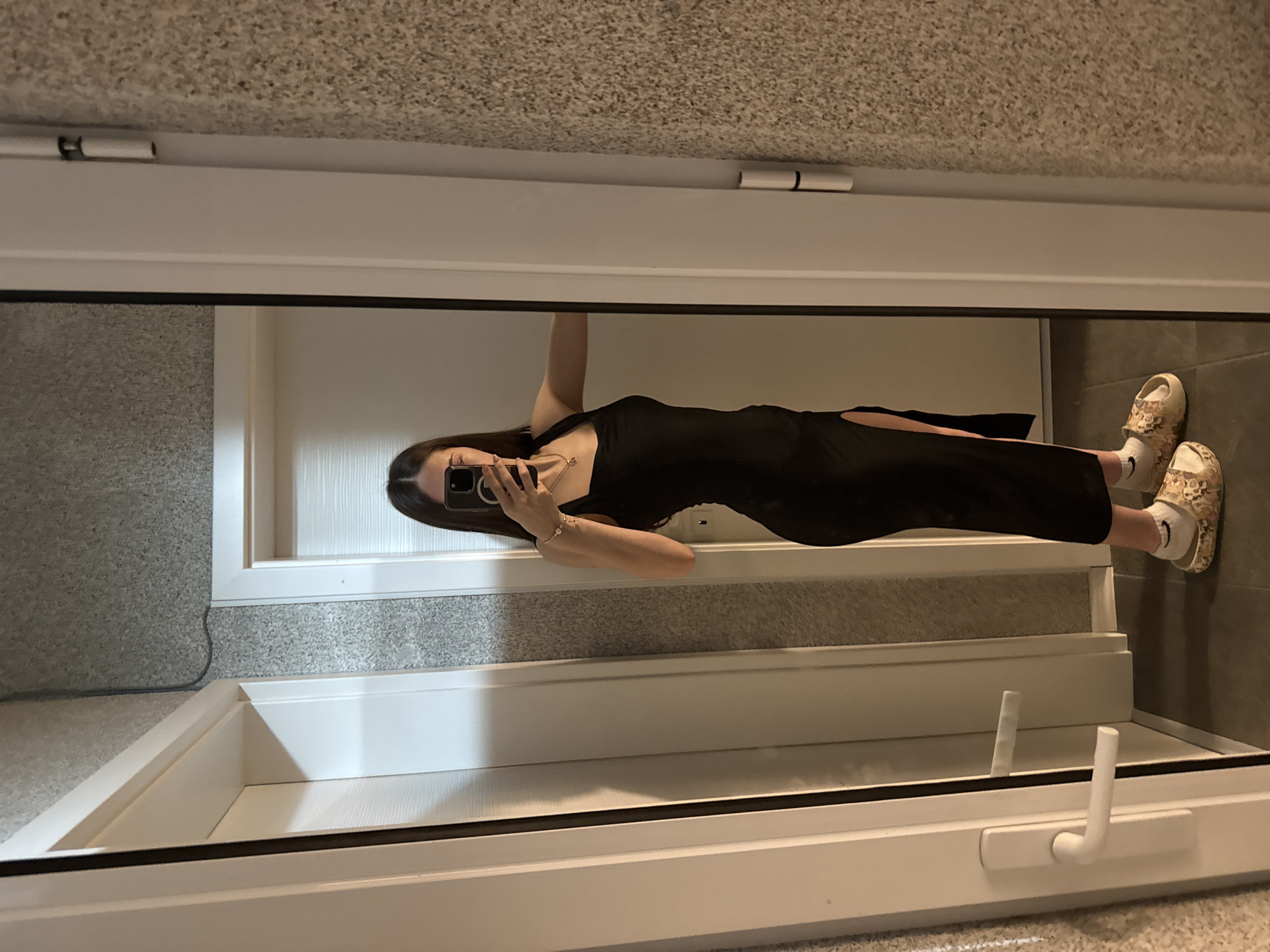 A woman is standing indoors, facing a mirror, and taking a selfie with her smartphone. She is wearing a black dress with a slit on the side and patterned clogs with white socks. The reflection shows a doorway and possibly a window frame. The room has tiled walls. The location is likely in Sofia, Bulgaria.
