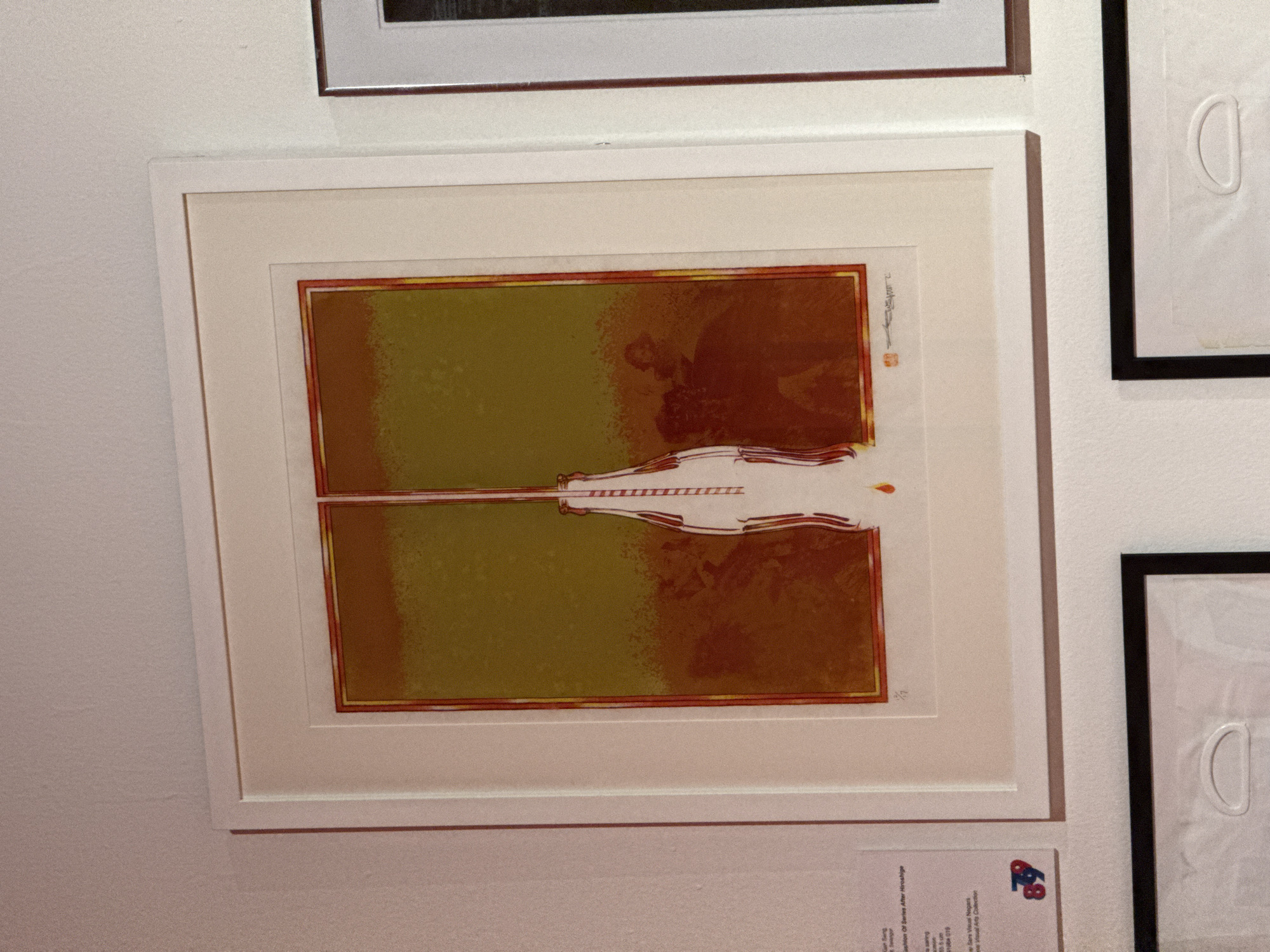 The image shows a framed artwork hanging on a white wall, likely in an art gallery or exhibition in Kuala Lumpur, Malaysia. The artwork is a print with a horizontal orientation, featuring an abstract image predominantly in shades of olive green and brown. A white, stylized depiction of what may be a train engine or stylized vehicle bisects the artwork horizontally. The print is double matted with a wider off-white mat and a thinner gold colored one. The frame is white. Additional framed artwork is partially visible above and to the right of the featured print. Below the artwork, there is a small information card with text and logos.