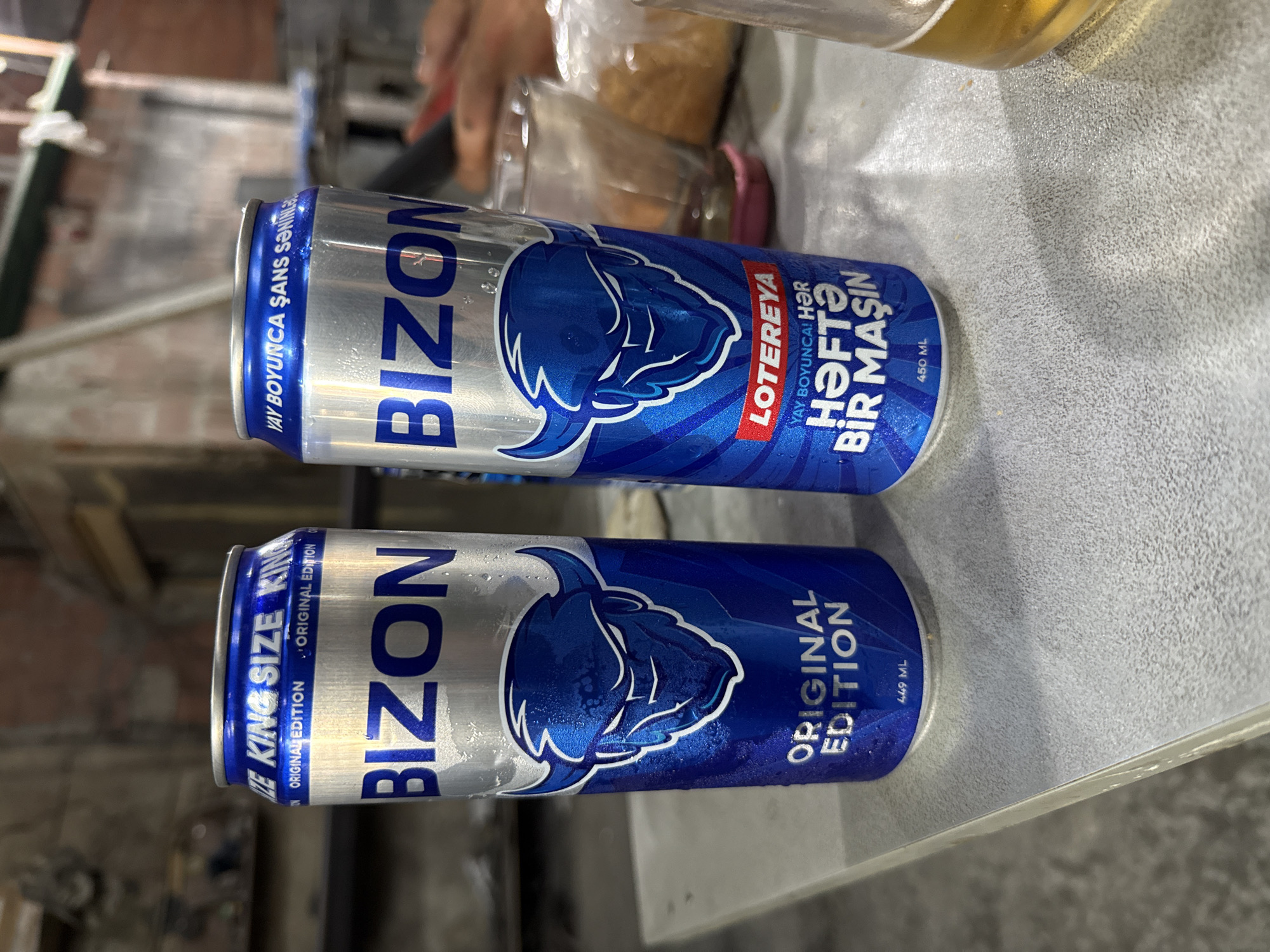 Two cans of Bizon energy drink are displayed on a gray, textured table. The can on the left is taller, with "KING SIZE" and "ORIGINAL EDITION" printed in blue on a silver background. The can on the right, shorter, also features "Bizon" and "ORIGINAL EDITION," along with a red "LOTEREYA" label and text that appears to be in Azerbaijani. Both cans show condensation and have a graphic of a blue bull's head. In the background, out of focus, are blurry figures of people, glasses, and what might be food items. The overall setting appears to be an indoor space with concrete walls, suggesting a casual or potentially industrial environment. The image was taken in Bilajari, Azerbaijan.