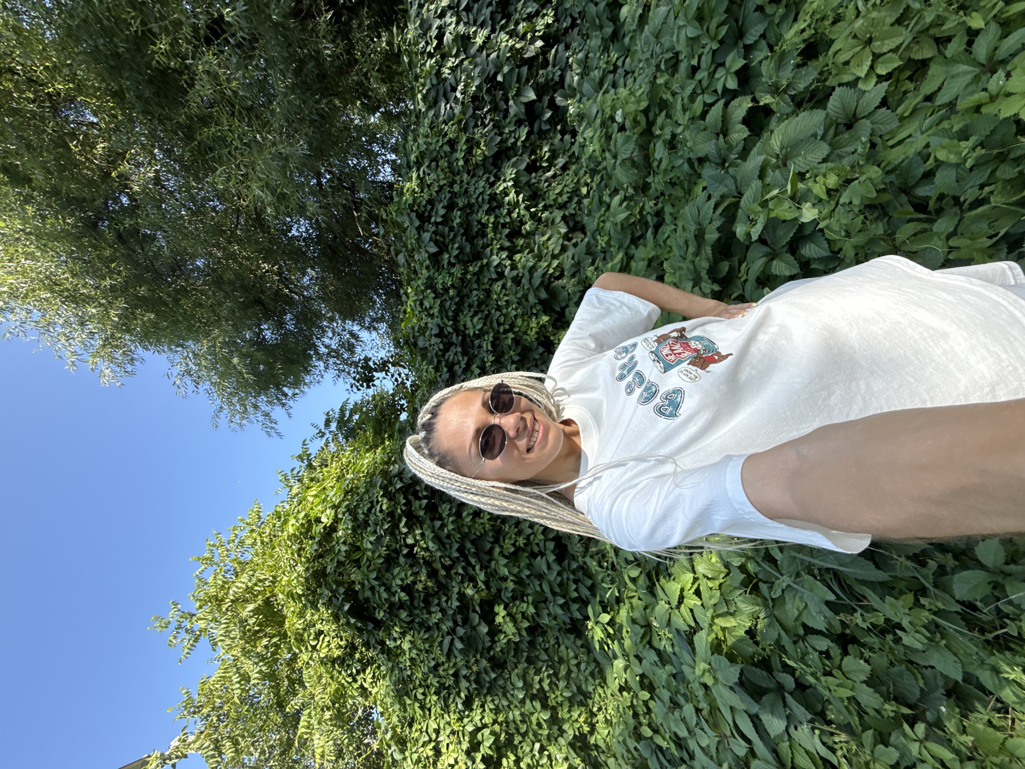 A young woman with blonde braided hair and sunglasses is taking a selfie outdoors. She is wearing a white t-shirt with a graphic design. She is standing in front of a dense green hedge, with trees and a blue sky in the background. The image appears to have been taken during daylight. The location is Rusanovo, Russia.